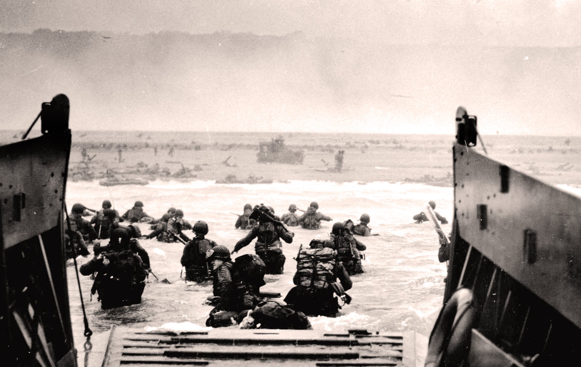 A black and white historical photo of soldiers disembarking from a landing craft onto Omaha Beach, France. The soldiers are carrying heavy backpacks and equipment. The photo is taken from the perspective of the landing craft, looking out towards the beach.