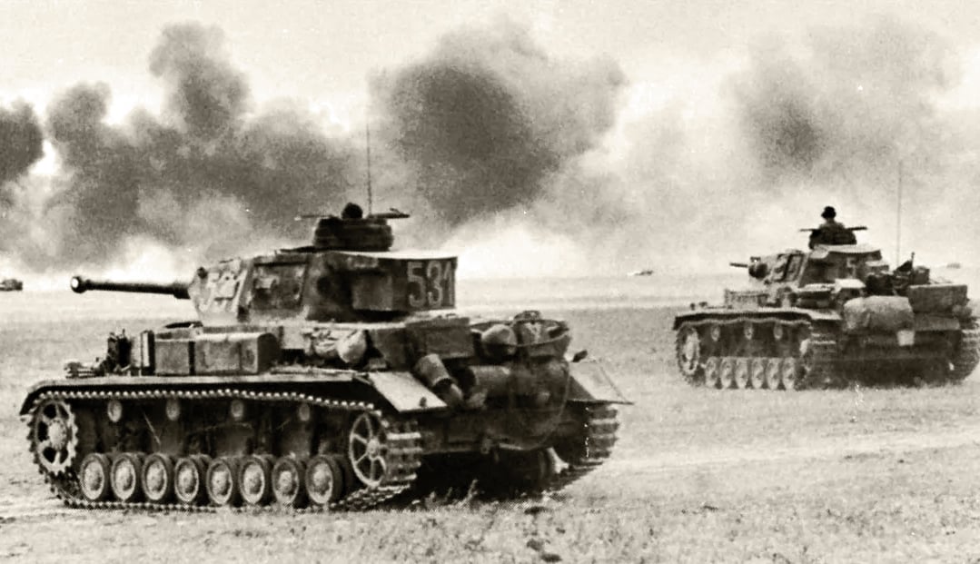 A black and white photo of two German Panzer Mark IV (foreground) and Panzer Mark III (background) tanks in a field with smoke rising from explosions in the background.