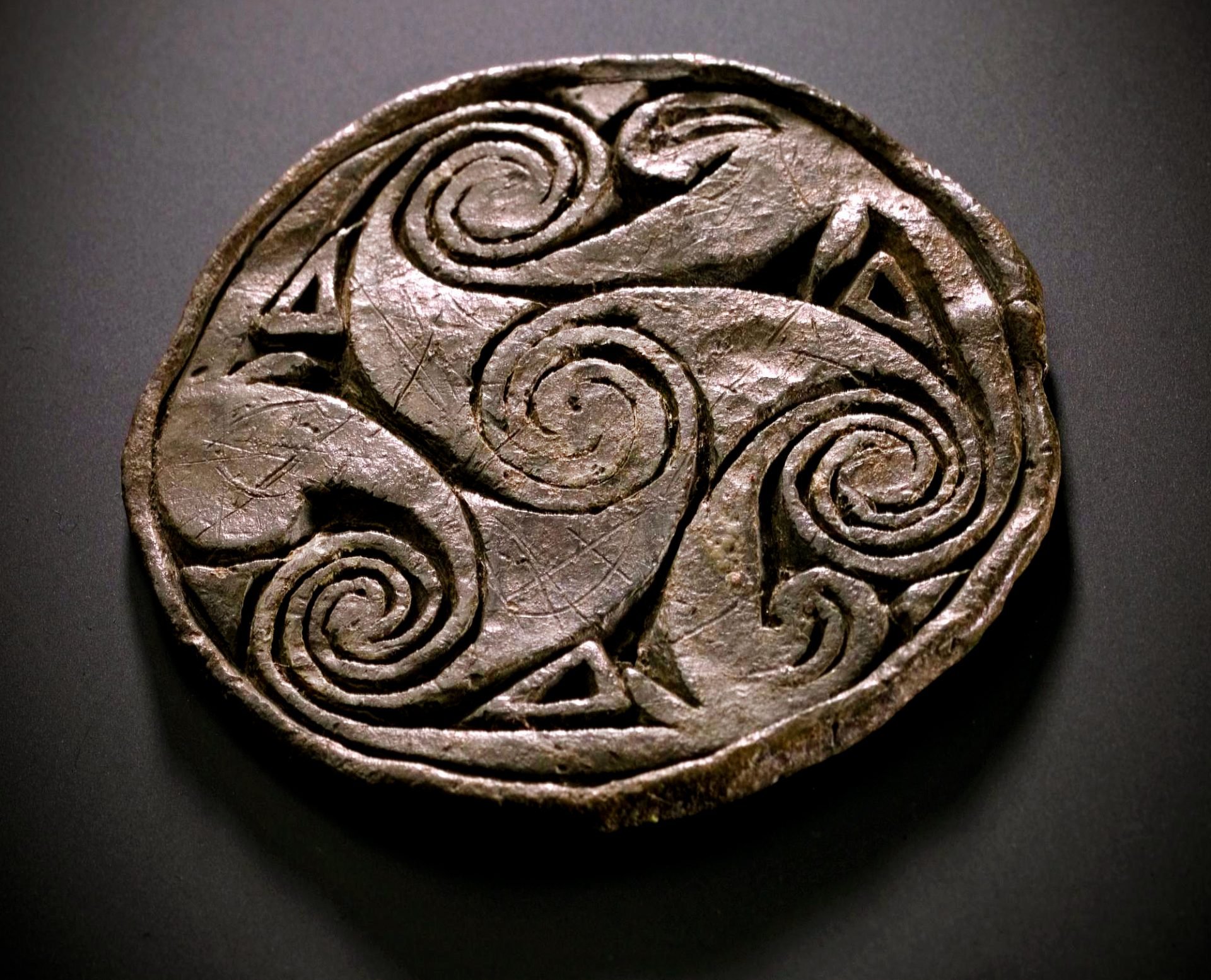 Pictish lead disc on a black background from the Brough of Birsay, Orkney, dated 700 - 900 AD, engraved with a Triskelion pattern and spirals.