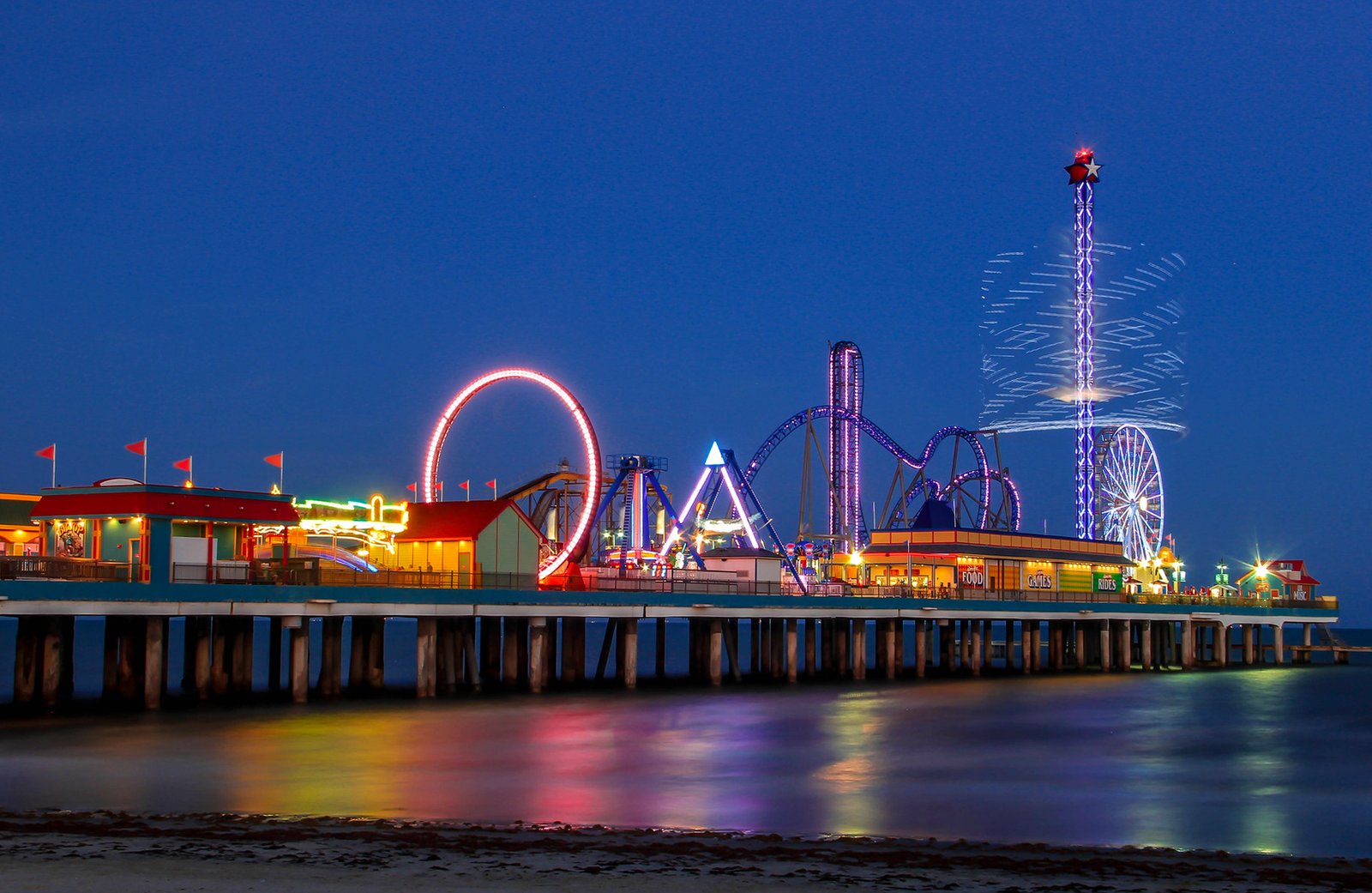 A night view of Pleasure Pier, an amusement park on a wooden pier in Galveston, Texas, with a roller coaster, a Ferris wheel, and a drop tower illuminated by colorful lights against a dark blue sky and ocean.