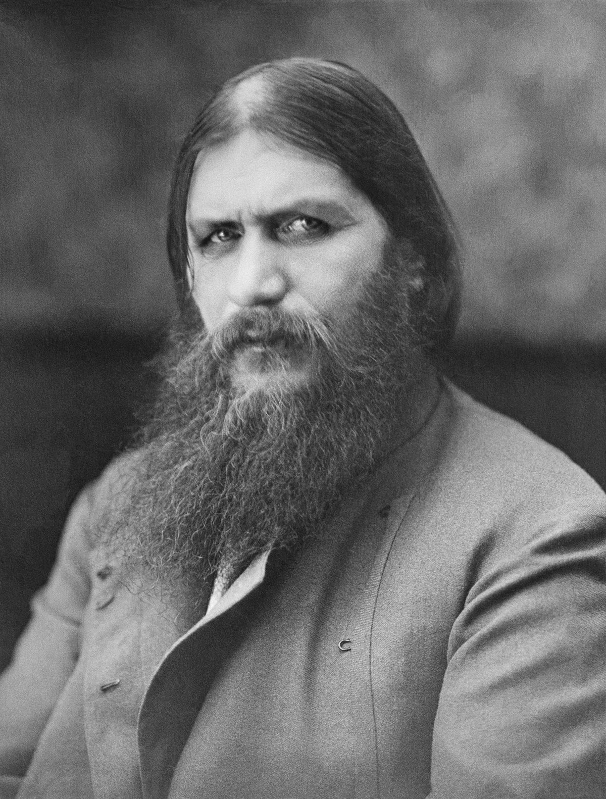 A portrait of Grigori Rasputin. He is looking into the camera. He has dark circles under his eyes and a long beard. He is wearing a simple gray jacket.