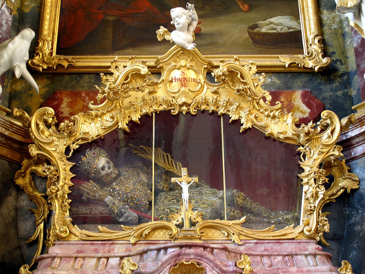 A photograph of a reliquary, a gilded elaborate box with a glass window. Lying inside is a skeleton of a saint dressed in golden clothing and a golden bejeweled crown.