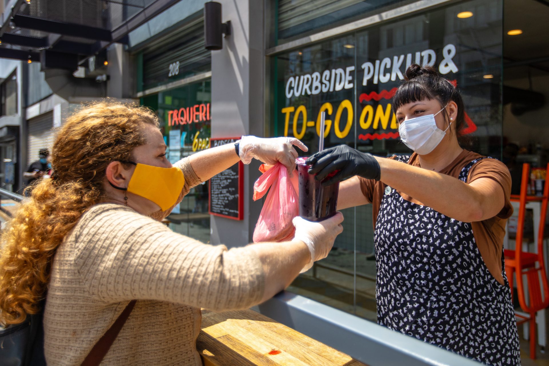 A photograph of a customer picking up takeout food outside of a restaurant. A restaurant employee is handing over a plastic bag and a soda cup to a customer. Both women are wearing face masks.