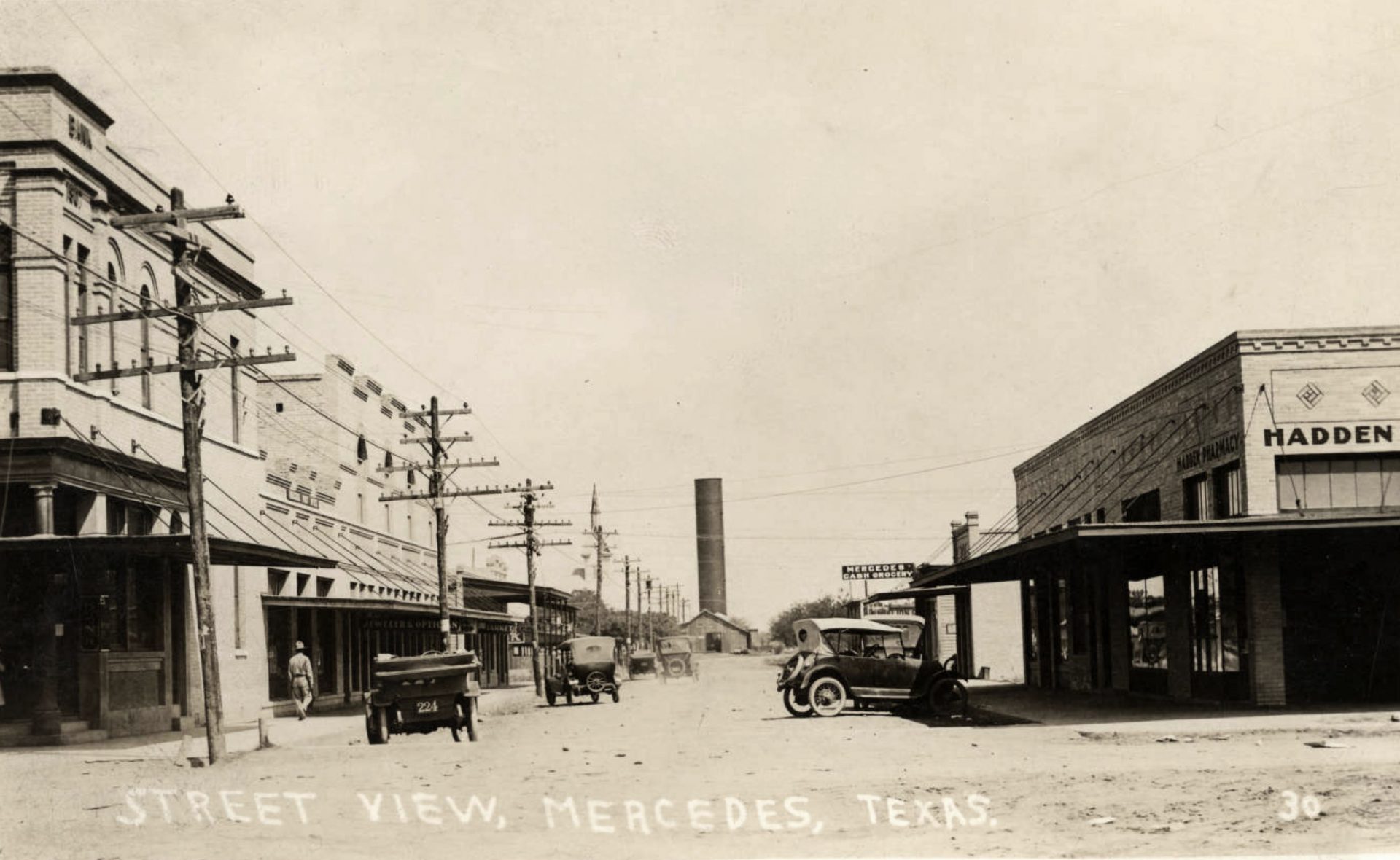 A black and white photograph of a street view in Mercedes, Texas, taken from the middle of the road. The street is lined with two-story buildings, some with signs or awnings. Cars are parked on the side of the road. A tall smokestack is visible in the background. The photograph has a caption that reads “Street View, Mercedes, Texas”.