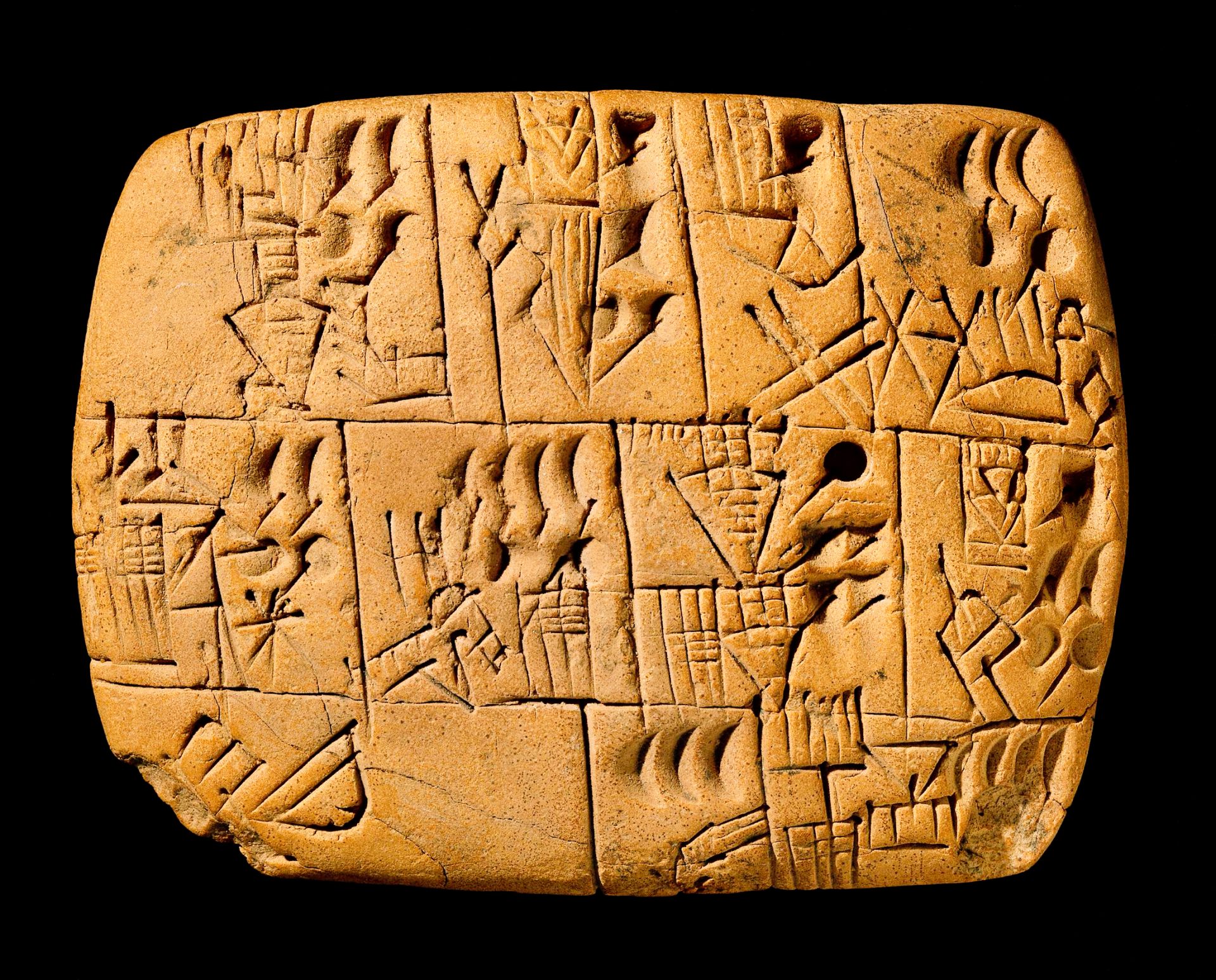 An ancient clay tablet with cuneiform writing on a black background. The tablet is rectangular in shape. The cuneiform writing is in a variety of sizes and styles, and covers the entire surface of the tablet. The tablet is light brown and has minor chips and cracks.