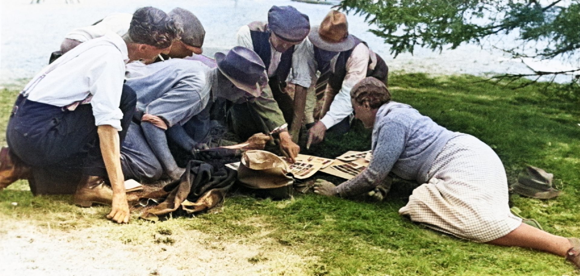 A group of people in old-fashioned clothing gathered around a stack of papers with photographs on a grassy area near Sutton Hoo.