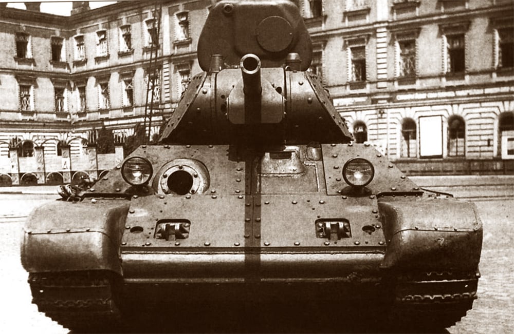 A black and white photo of a Soviet T-34 tank facing the camera in a city square with arched buildings in the background.