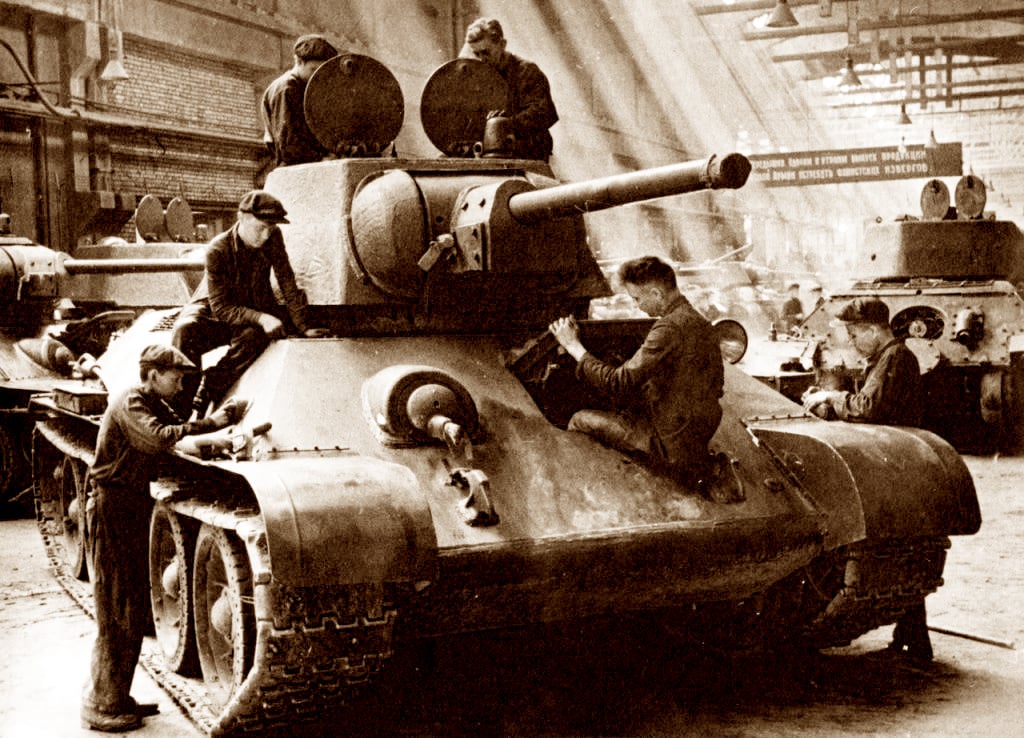 A black and white photo of a group of men working on a large tank T-34 assembly with a turret and a long barrel in a factory with other tanks and machinery in the background.