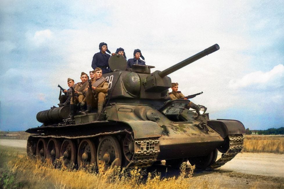 A historical photo of a group of Soviet World War II soldiers on a green T-34 tank with the number 28 in a field with trees and a blue sky.