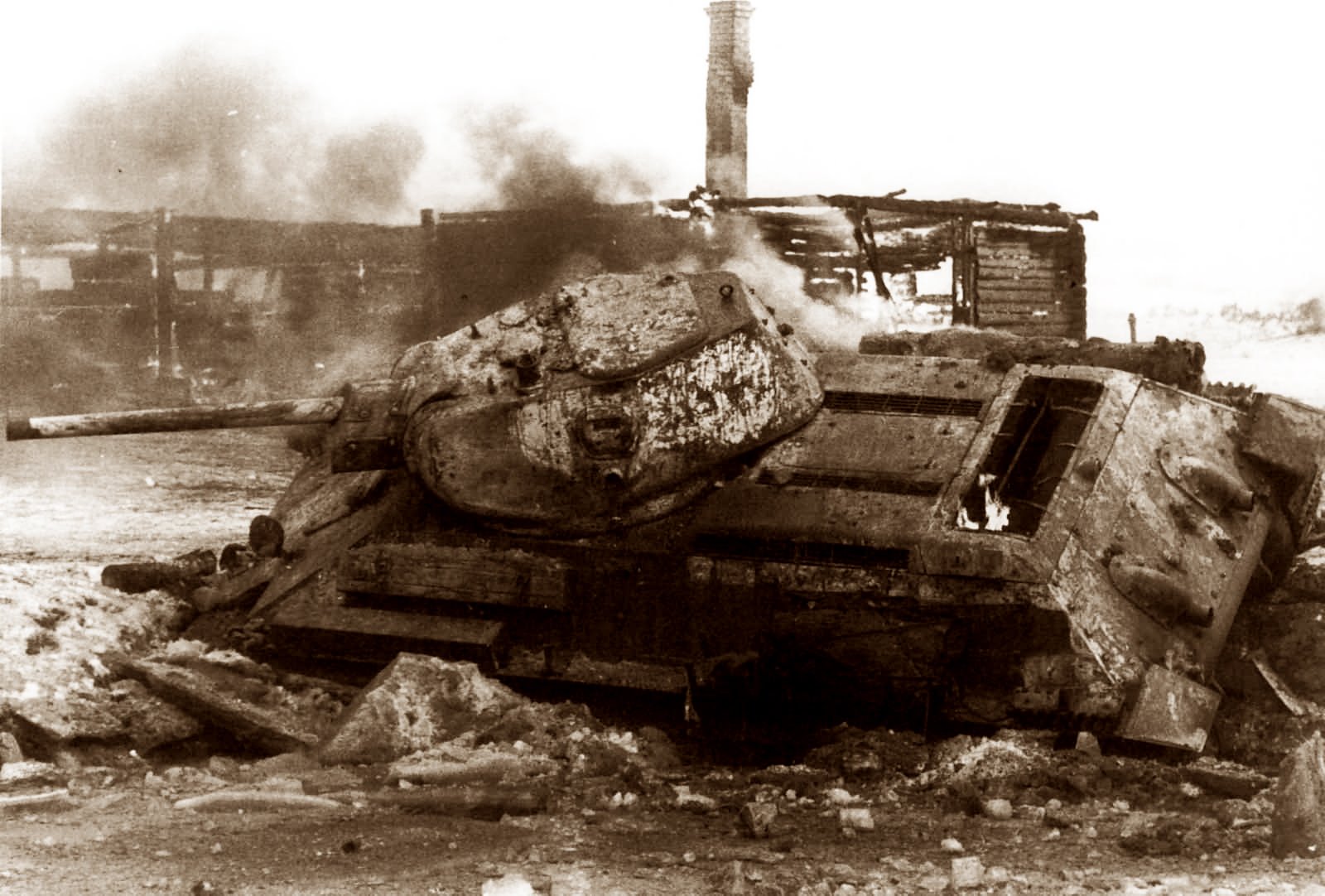 A black and white photo of a destroyed T-34 tank in a warzone with a burning building and a smokestack in the background.