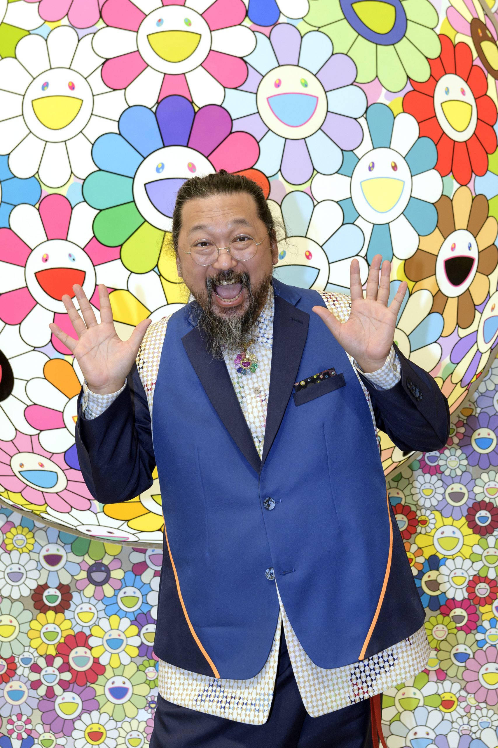 A photograph of Takashi Murakami, a Japanese man with his mouth open and his hands raised in the air in a playful manner. He is wearing a long blue jacket. The background features smiling flowers in various colors.