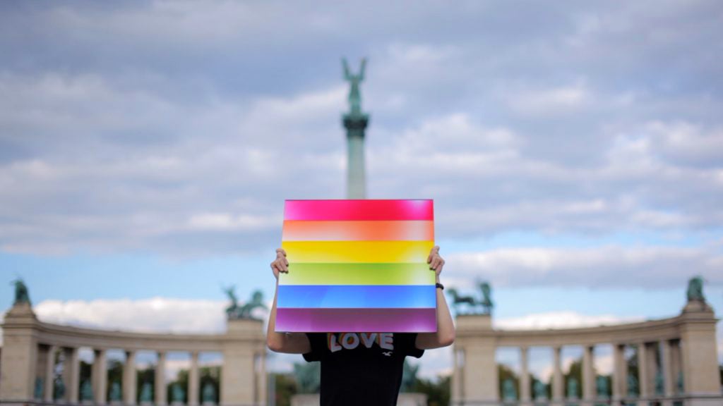 A photo realistic image of a person holding a rainbow flag in front of a monument, expressing support for LGBTQ+ rights. The person is wearing a black t-shirt with the word “LOVE” written on it, matching the message of the flag. The rainbow flag is made up of horizontal stripes of different colors - red, orange, yellow, green, blue, and purple - representing the diversity and inclusion of the LGBTQ+ community. The monument in the background is a tall column with a statue on top, symbolizing the history and culture of the place. It is surrounded by a colonnade, creating a contrast between the classical and modern styles.
