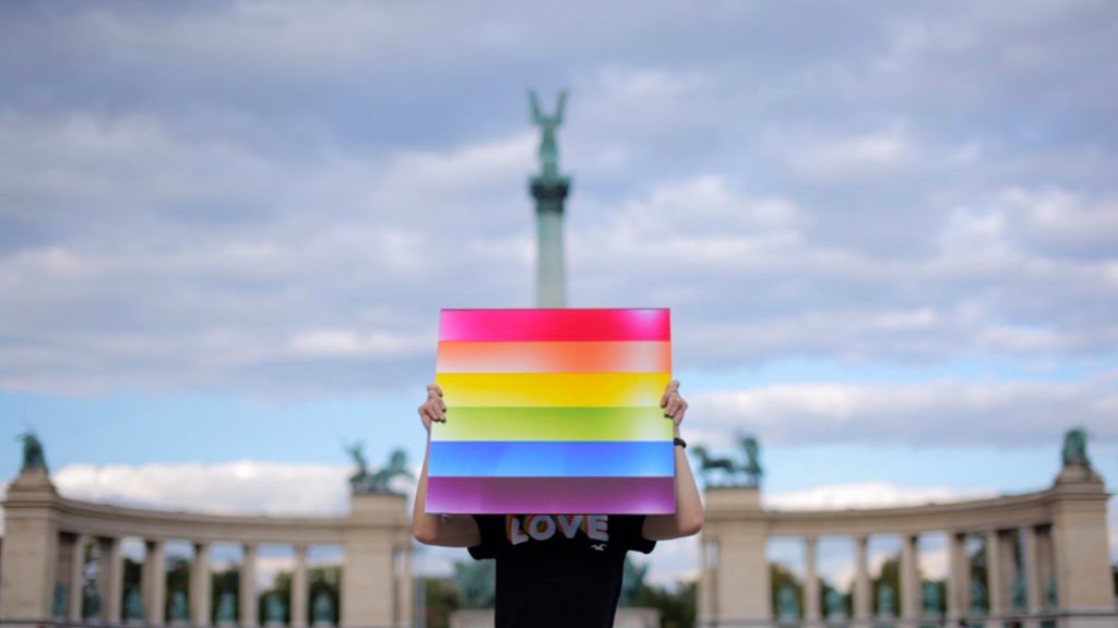 A photo realistic image of a person holding a rainbow flag in front of a monument, expressing support for LGBTQ+ rights. The person is wearing a black t-shirt with the word “LOVE” written on it, matching the message of the flag. The rainbow flag is made up of horizontal stripes of different colors - red, orange, yellow, green, blue, and purple - representing the diversity and inclusion of the LGBTQ+ community. The monument in the background is a tall column with a statue on top, symbolizing the history and culture of the place. It is surrounded by a colonnade, creating a contrast between the classical and modern styles.