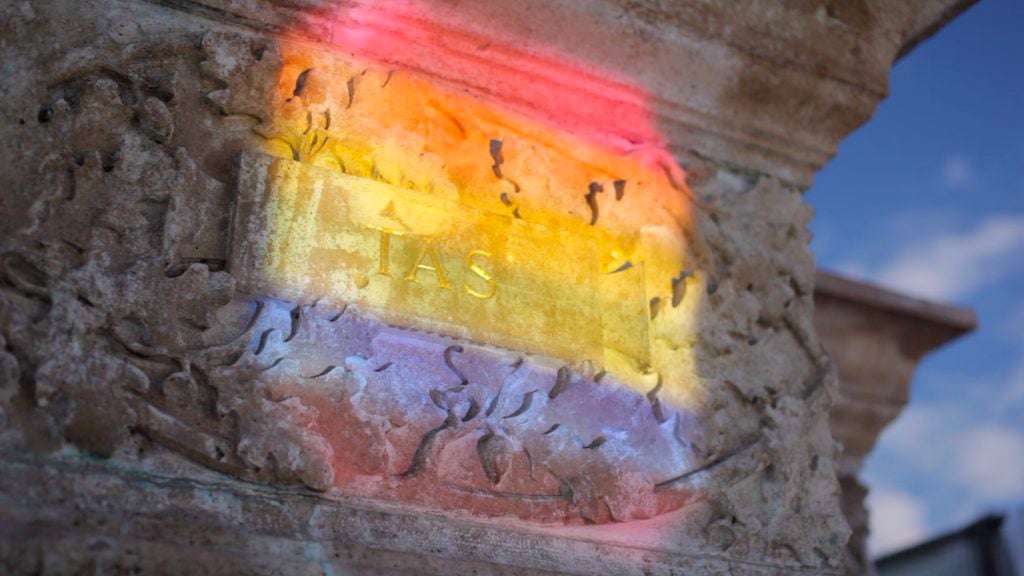 A photo of a stone architectural detail with a rainbow of colors projected onto it, creating a striking visual effect. The stone detail is a rectangular block with a carved border and the word “TAS” in the center. The colors projected onto the stone are pink, orange, yellow, green, blue, and purple, representing the diversity and pride of the LGBTQ+ community. The photo appears to be taken from below, looking up at the stone detail, giving it a sense of prominence and importance.