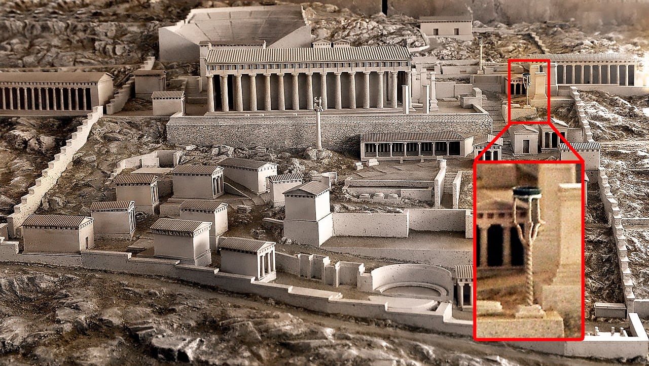 A photograph showing a miniature replica of Delphi's ancient cityscape, set against a backdrop of hilly terrain. A red rectangle emphasizes where the cauldron once stood.