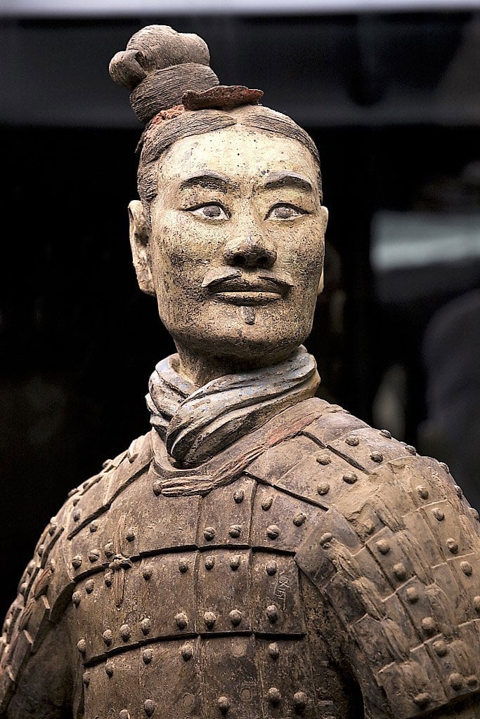 A photograph of a terracotta warrior clay statue. The warrior is wearing full armor, his hair is tied up into a bun.