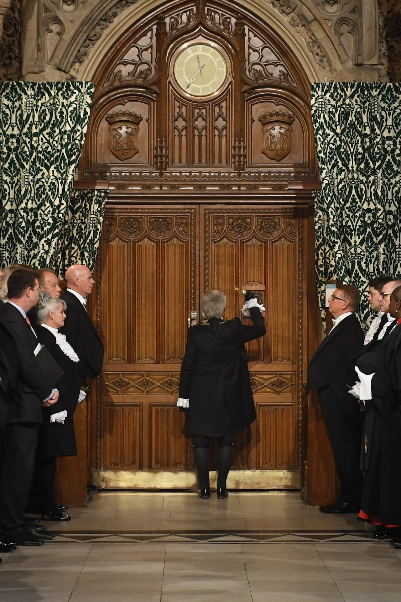 A photograph of a person dressed in black knocking on a large wooden door.