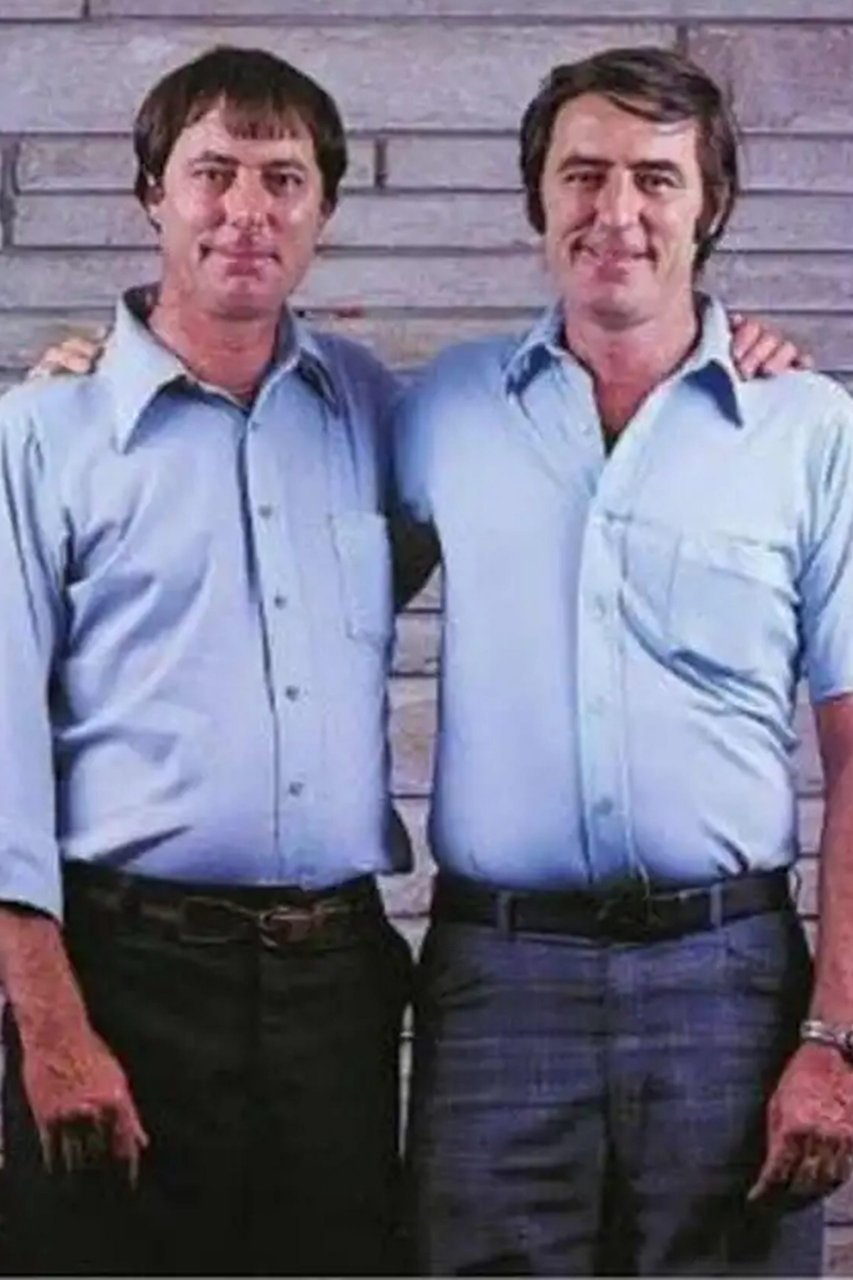 A photo of two male identical twins standing together. Both are wearing blue shirts and dark colored pants.
