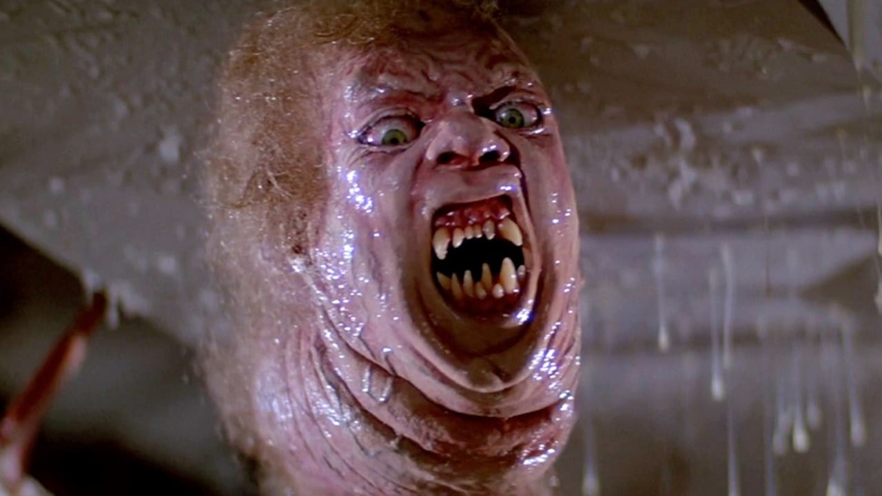 A still image from the film The Thing, featuring a worm like monster with a slimy humanoid head and sharp teeth.