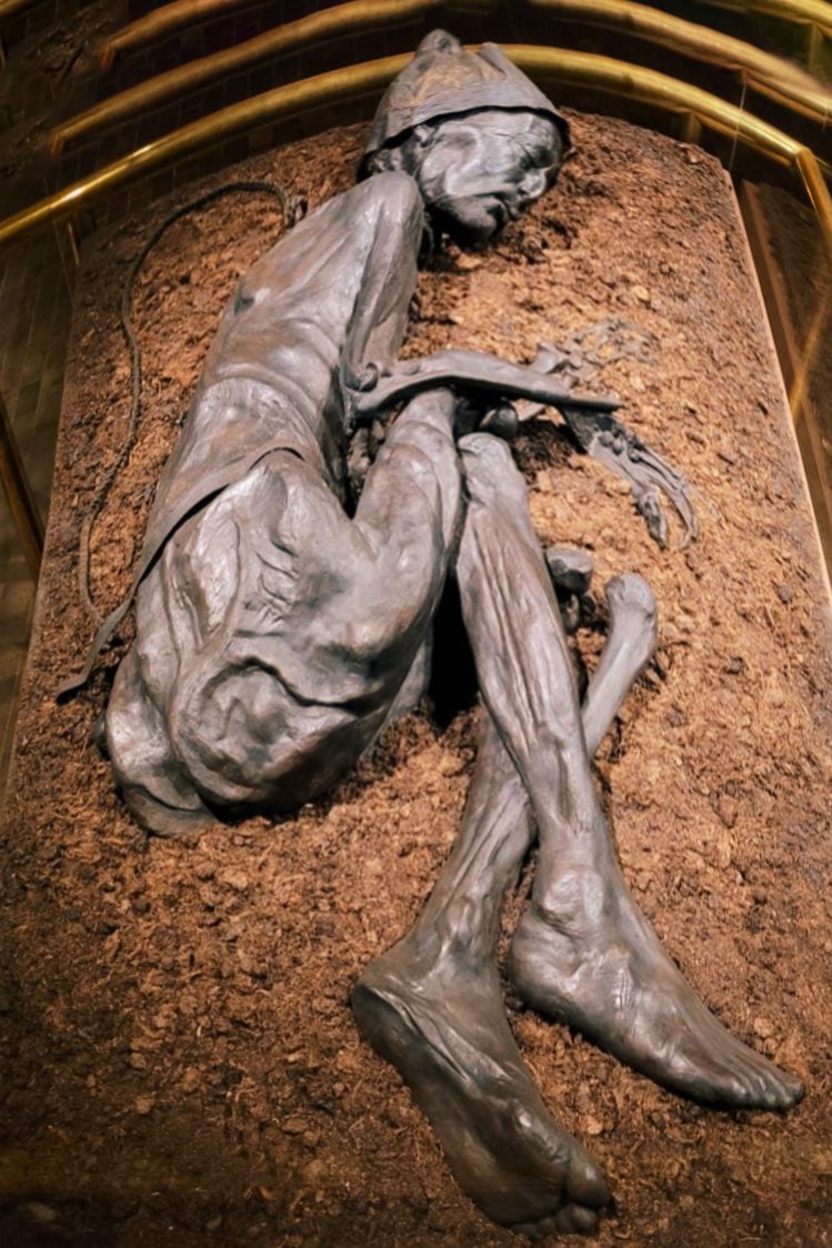 A photograph of a mummified man in the fetal position. The body has a hat on it's head and is dark brown in color.