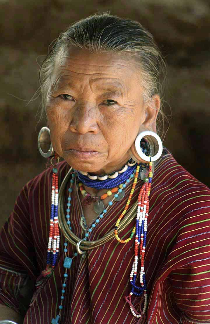 A photograph of an elderly Kayaw tribe woman. She is wearing traditional jewelry and large, ear expanding metal circle earrings. She is looking straight at the camera with her eyes wide open.