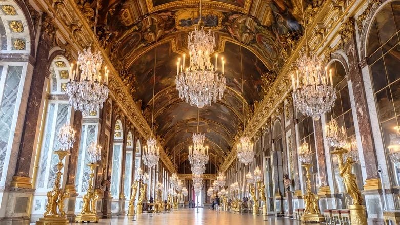 Photograph of The Hall of Mirrors at Chateau Versailles showcasing crystal chandeliers, golden decor and floor to ceiling mirrors.