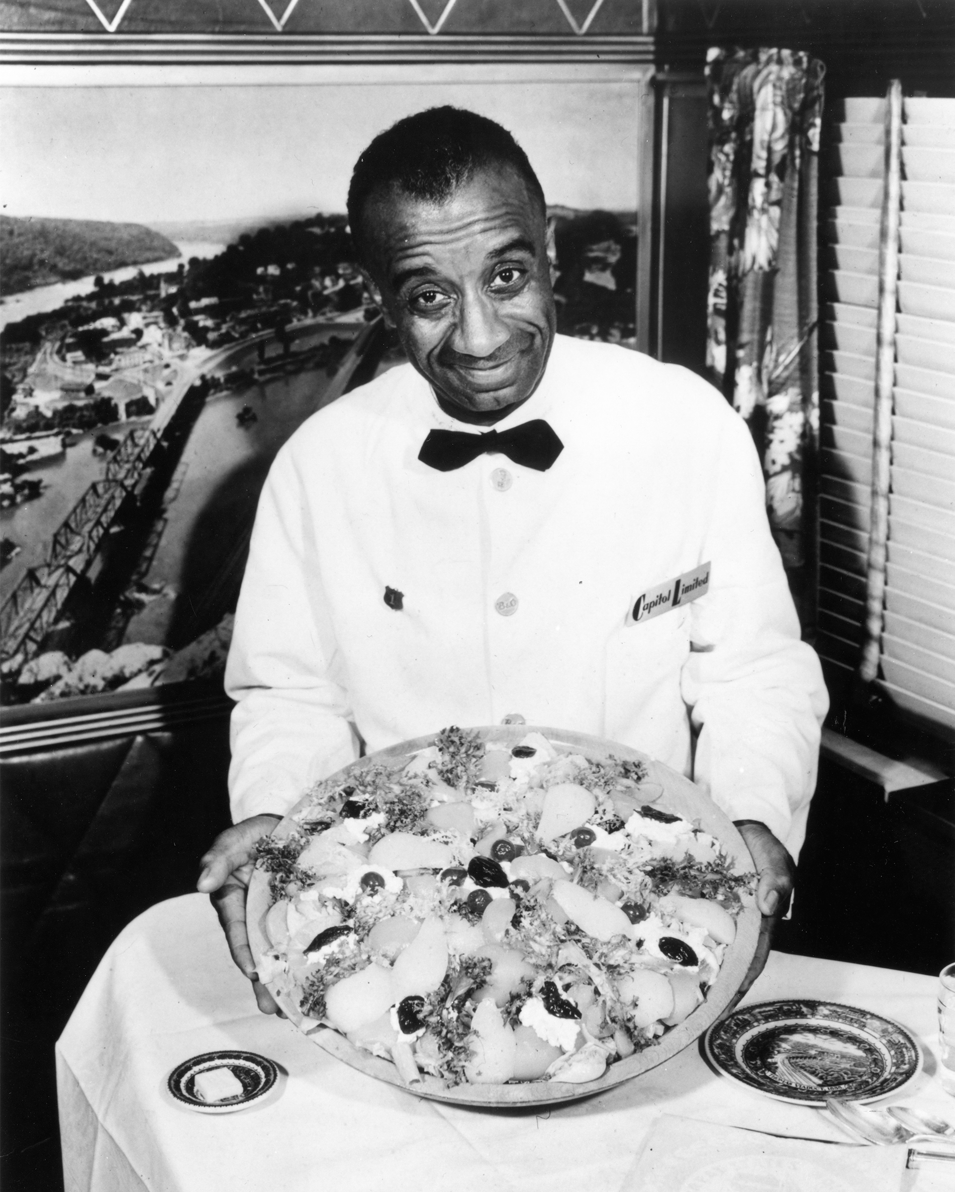 This black and white photograph shows a man in a white waiter's uniform and a black bow tie holding a large plate of food above a table. The plate has several different items on it.