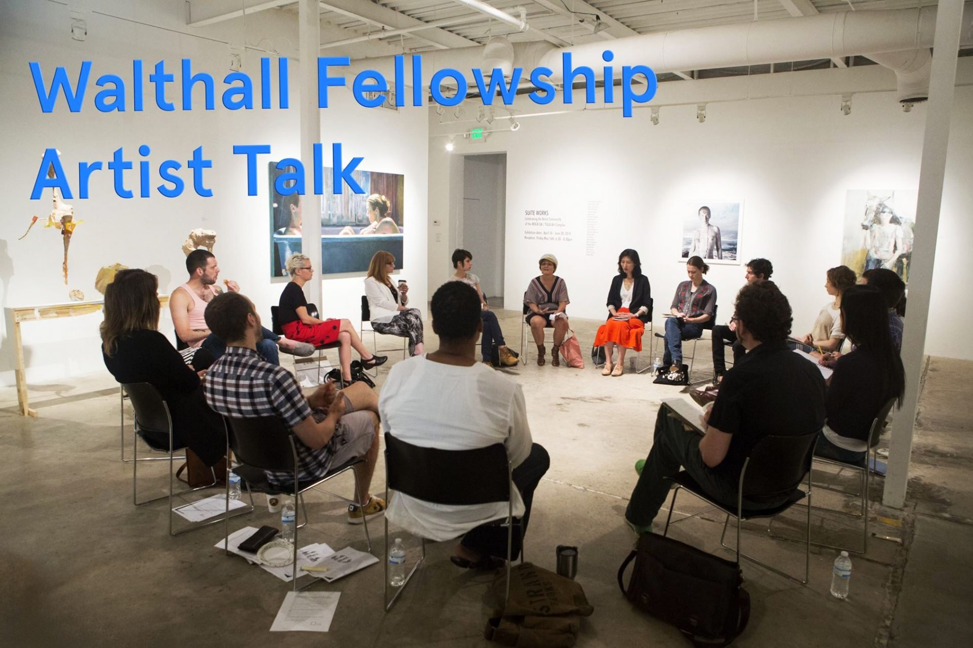 An artist talk event in a gallery space with the title “Walthall Fellowship Artist Talk” on the wall. Several people are sitting in a circle on folding chairs and talking. The gallery has white walls and concrete floors, with sculptures and artwork on display.