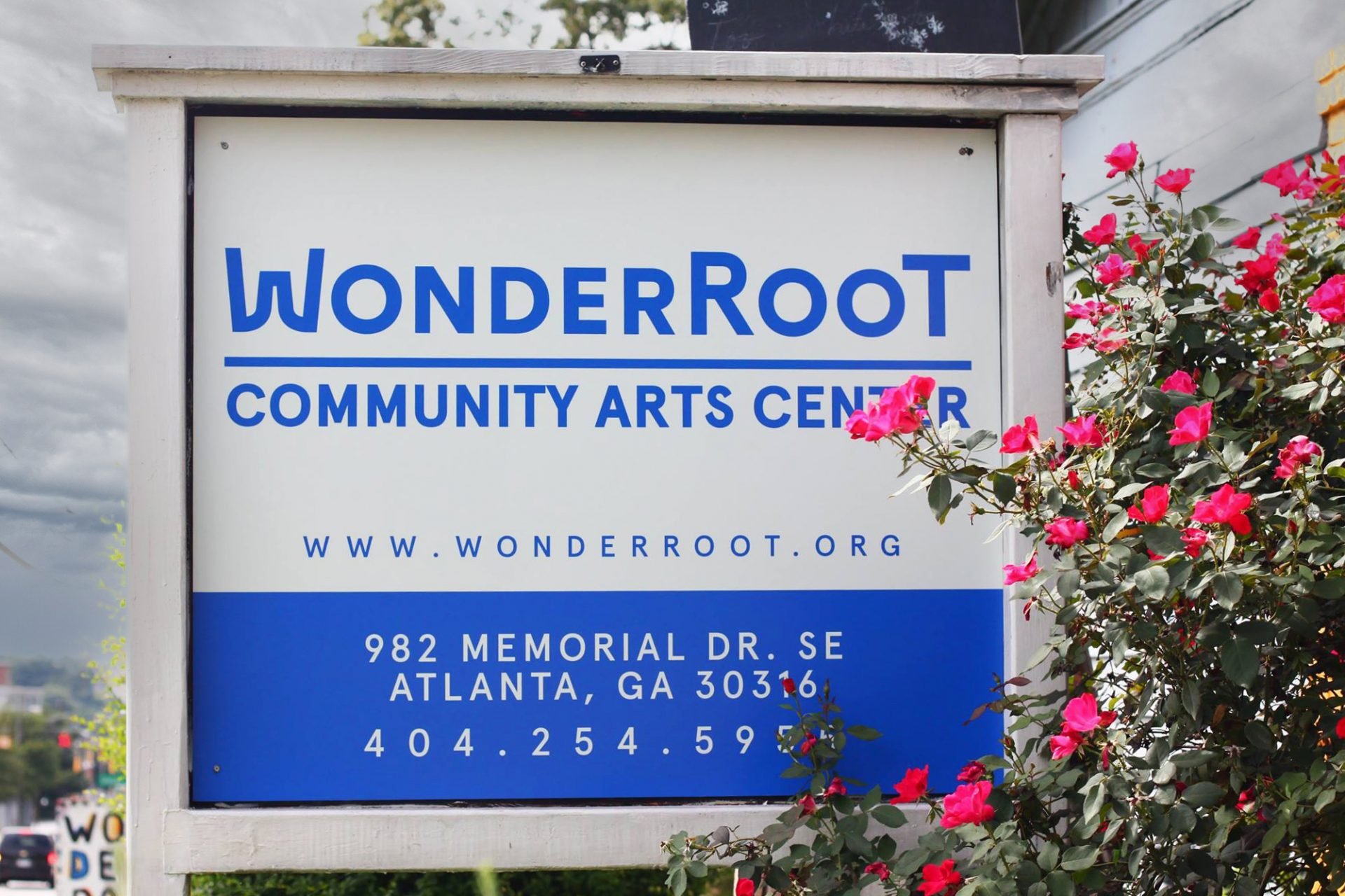 A sign for the WonderRoot Community Arts Center with a white background and blue text that gives the name, website, and address of the center. The sign is framed by pink flowers and green leaves. The sky behind the sign is cloudy.