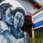 A photo collage of a mural of two people hugging and a photo of a building with a red roof and a sign that says “WonderRoot Community Arts Center”. The building is surrounded by trees and bushes with colorful autumn leaves.