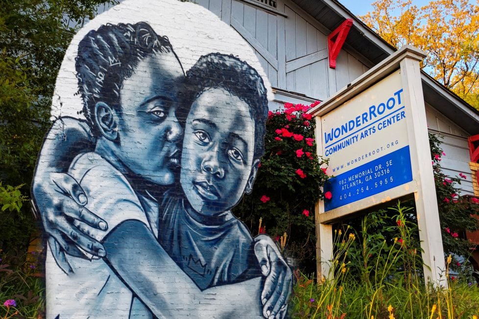 A photo collage of a mural of two people hugging and a photo of a building with a red roof and a sign that says “WonderRoot Community Arts Center”. The building is surrounded by trees and bushes with colorful autumn leaves.