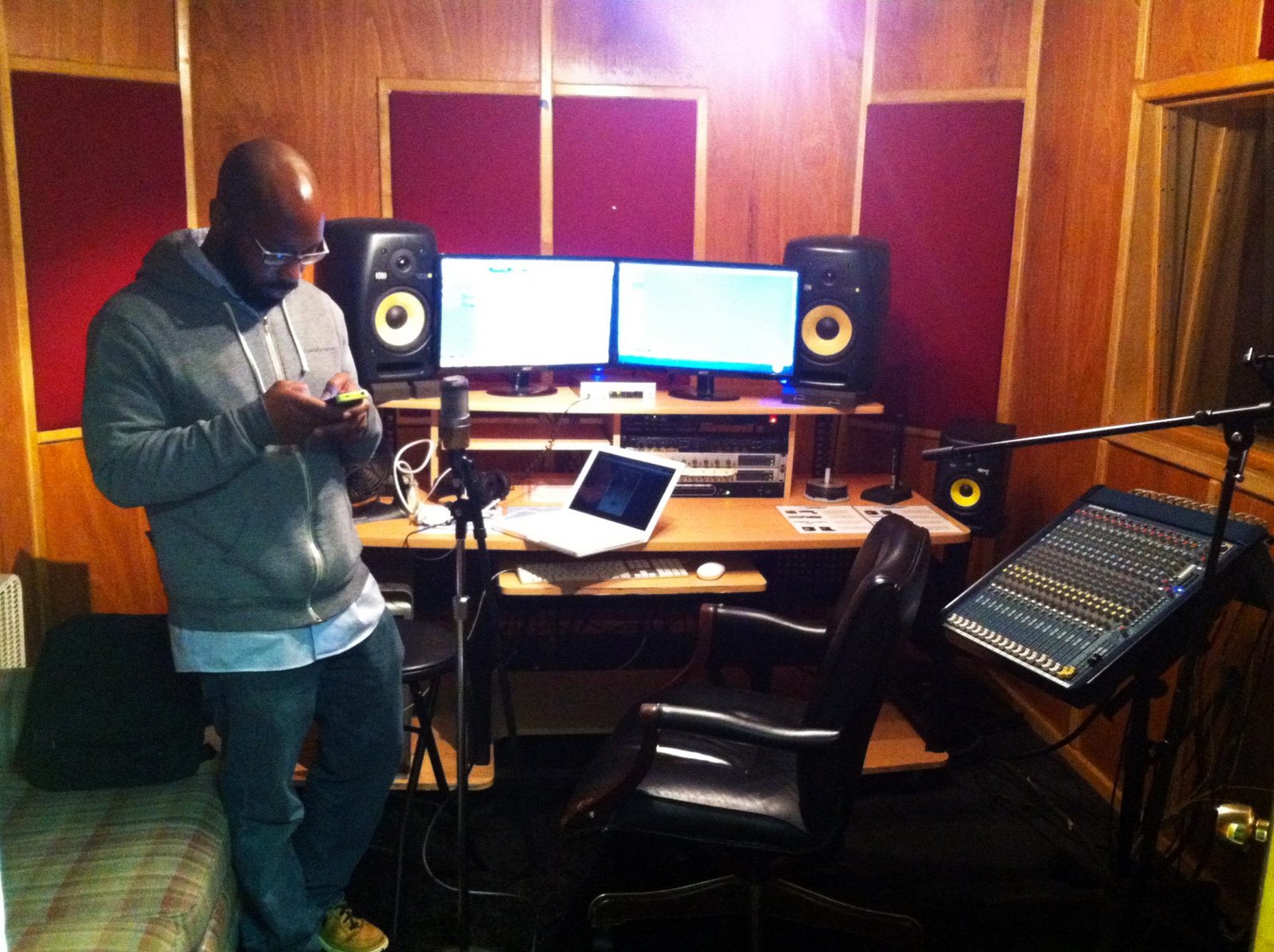 A man is standing in front of a microphone in a recording studio. The studio has a desk with multiple computer monitors and speakers, a mixing console, and wood paneling on the walls.