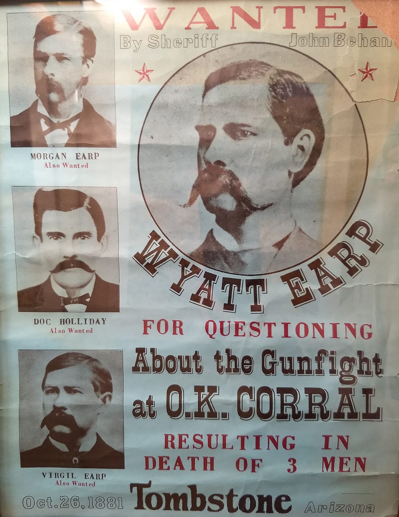 The poster shows a mugshot of four men, the man in the center of the poster has a mustache standing in front of a large white wall. The man is wearing a dark suit and has a look of determination on his face. The word "Wanted" written in black text at the top.