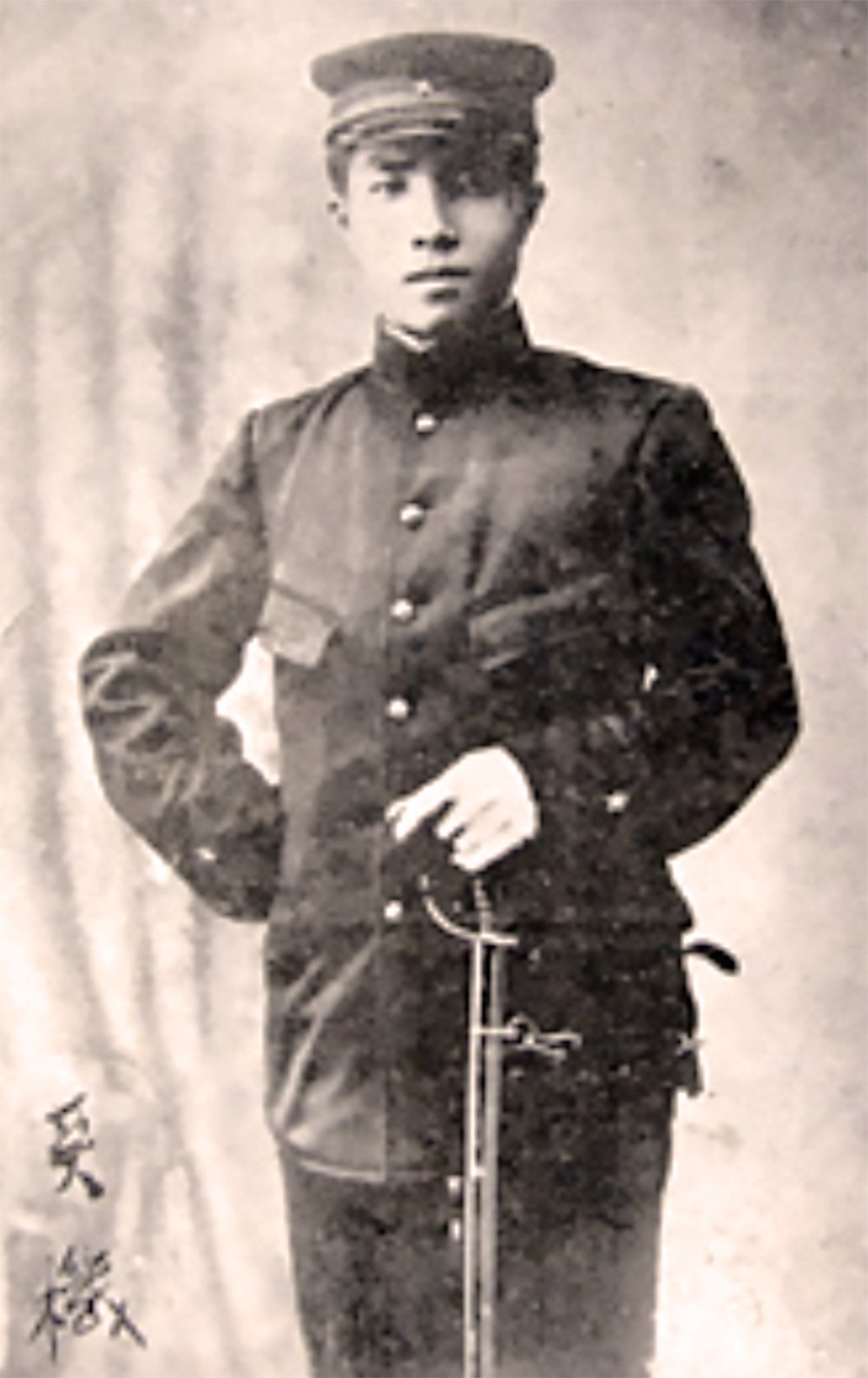 A historical photo of young Hideki Tojo in a military uniform holding a sword. He is standing with a confident pose and is wearing a military cap. The background is light-colored with a faint pattern.