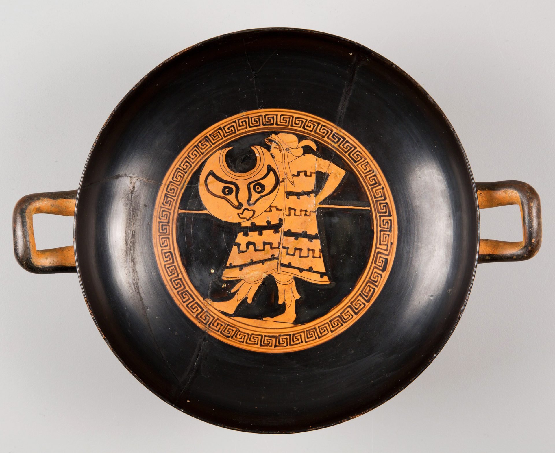 Black and orange painted ancient Greek kylix, or shallow drinking cup, with two handles. The central design features a detailed depiction of a warrior in Thracian attire, surrounded by a circular Greek key pattern.