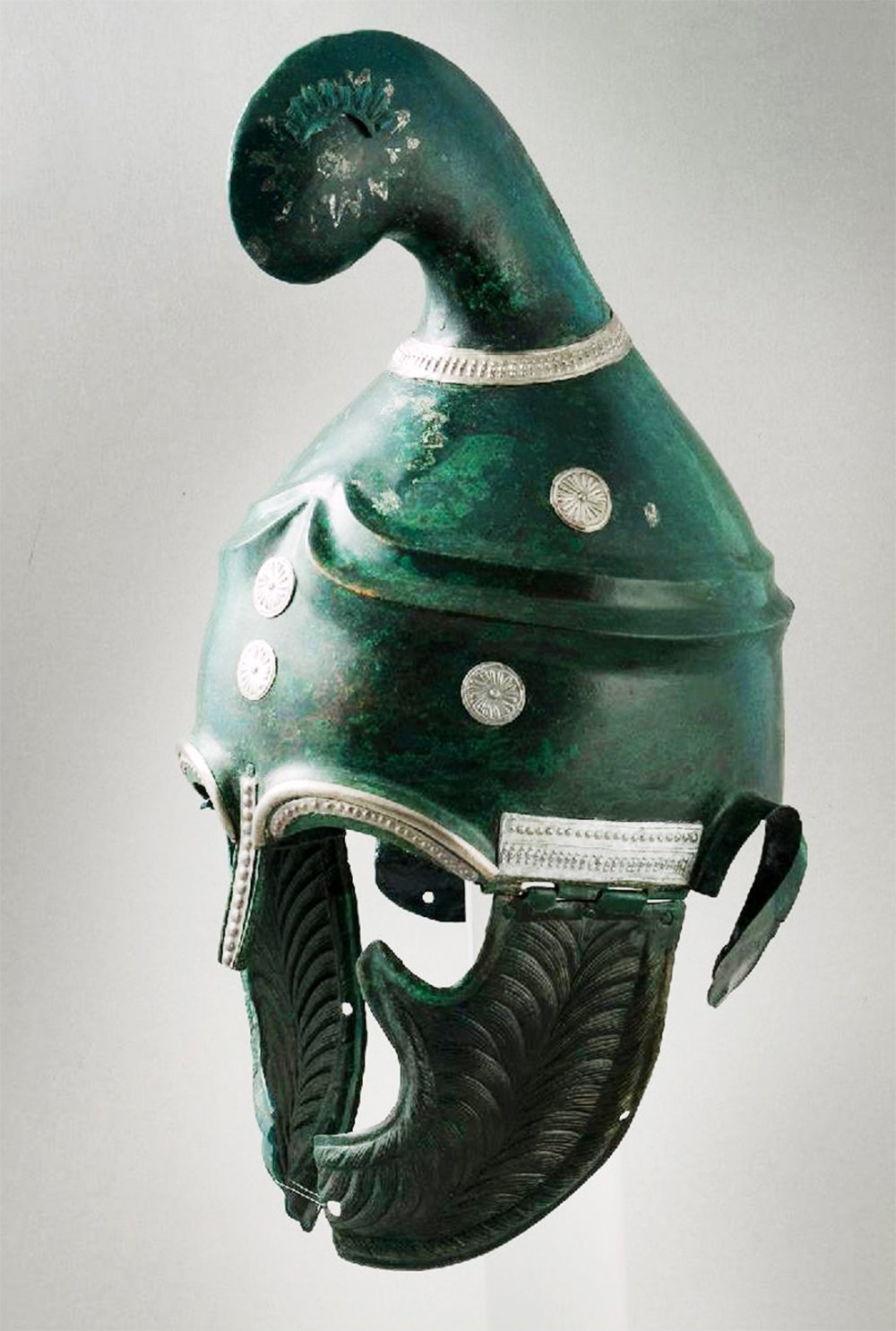Close-up of a 4th century BC Thracian-type helmet made of greenish-bronze, adorned with intricate silver detailing. The helmet features a tall, curved crest on top and ornate feather-like cheek guards. Decorative round medallions and silvered edging add to the helmet's elegance.