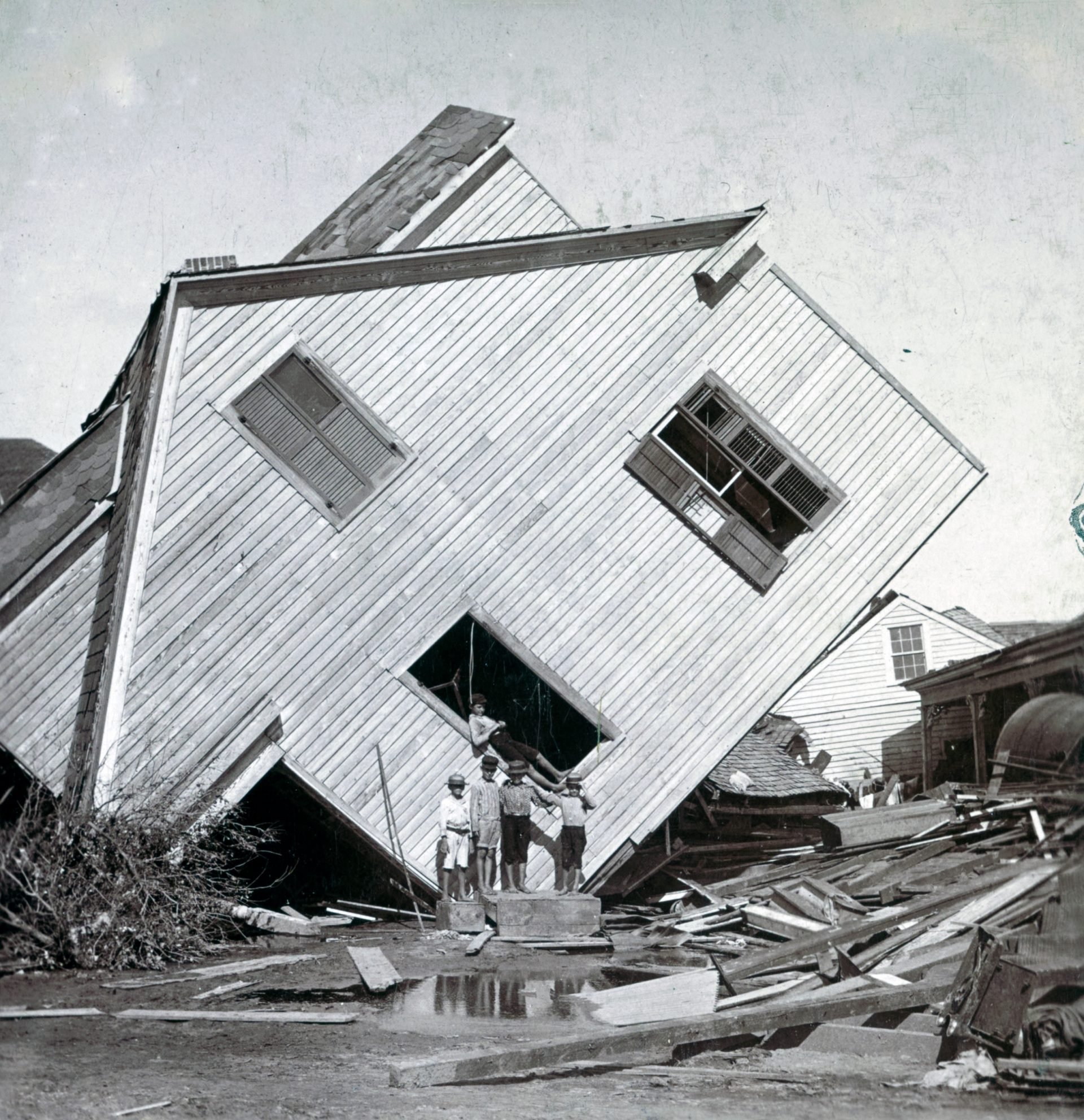 Photograph shows a house tipped on side, with several boys standing in front, after a flood in Galveston, Texas