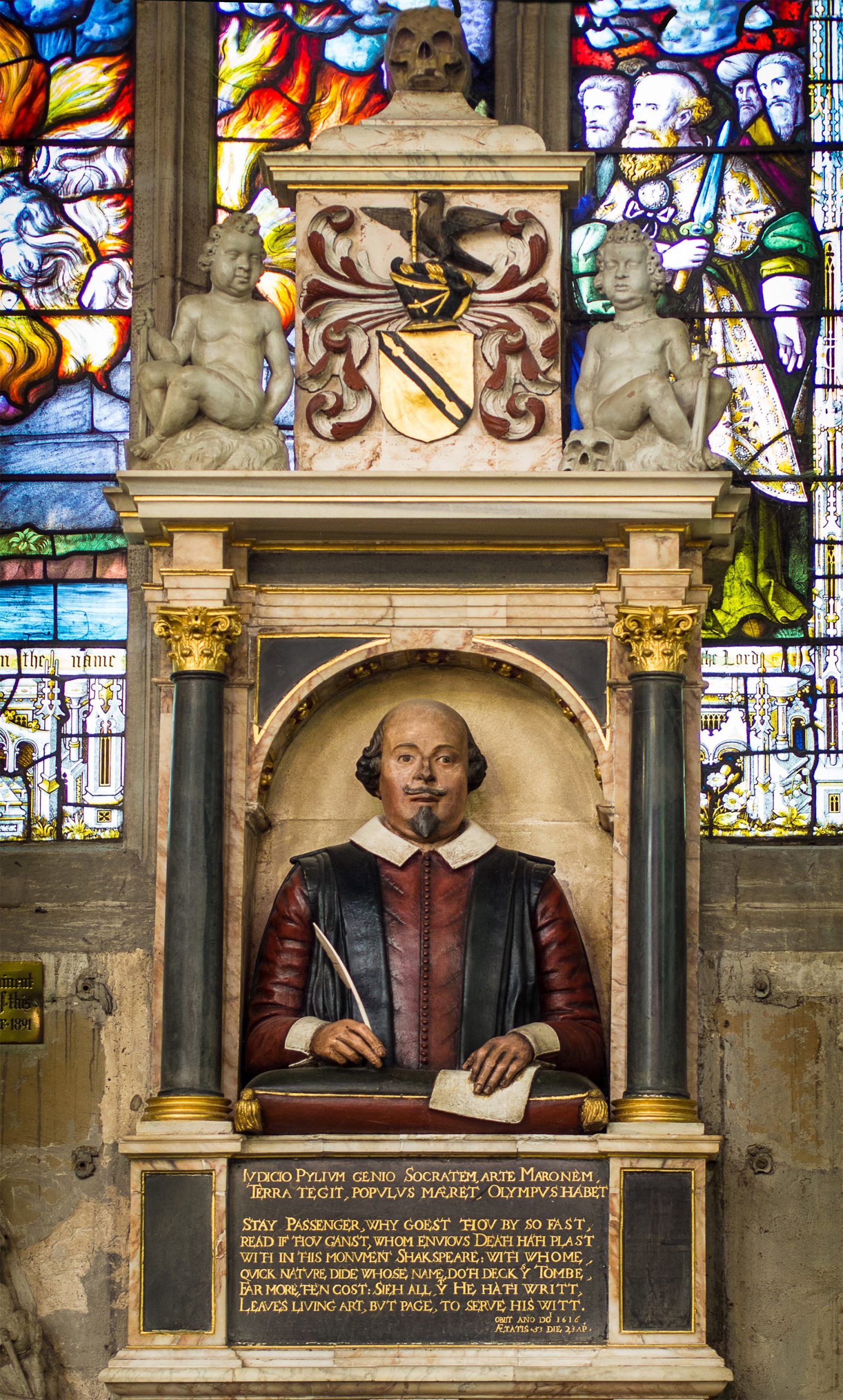 A grand monument inside Holy Trinity Church, Stratford, dedicated to William Shakespeare. The centerpiece is a life-like bust of Shakespeare, showing him holding a quill, with a focused expression. He is dressed in a red vest with a white collar. Above him, there are decorative elements, including a crest with a bird and a shield, flanked by two stone cherubs. Vibrant stained glass windows provide a colorful backdrop, illustrating religious scenes. Below Shakespeare's bust, an inscribed plaque with eloquent words pays homage to the playwright's legacy. The monument's intricate details and stonework highlight its historical and artistic significance.