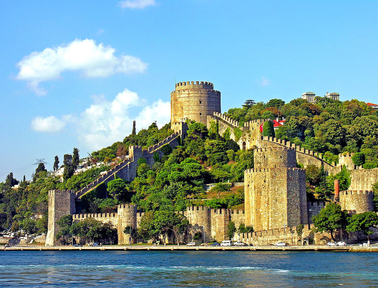 The majestic Rumeli Hisari Fortress situated on the European banks of the Bosphorus in Istanbul, Turkey. The fortress, made of stone walls and cylindrical towers, stretches across a lush hillside overlooking the sparkling waters of the Bosphorus. Green trees envelop the historical structure, and the clear blue sky above is dotted with a few white clouds. In the foreground, the serene waters reflect the stone walls of the fortress. This historic site stands as a testament to Istanbul's rich heritage.