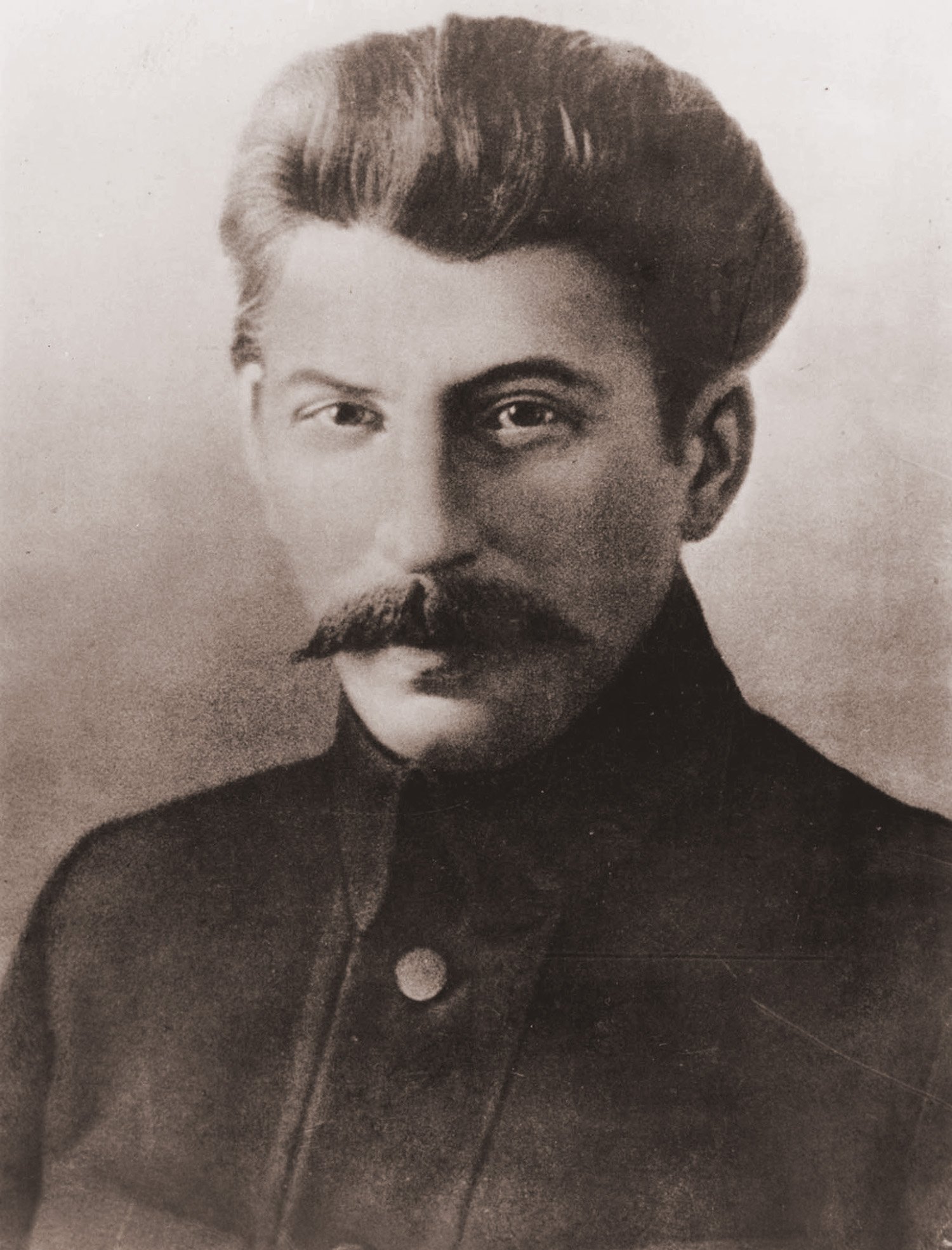 Sepia-toned portrait of a young Joseph Stalin from 1917, featuring his distinct mustache and a prominent swept-back hairstyle. He is wearing a dark jacket with a buttoned collar.
