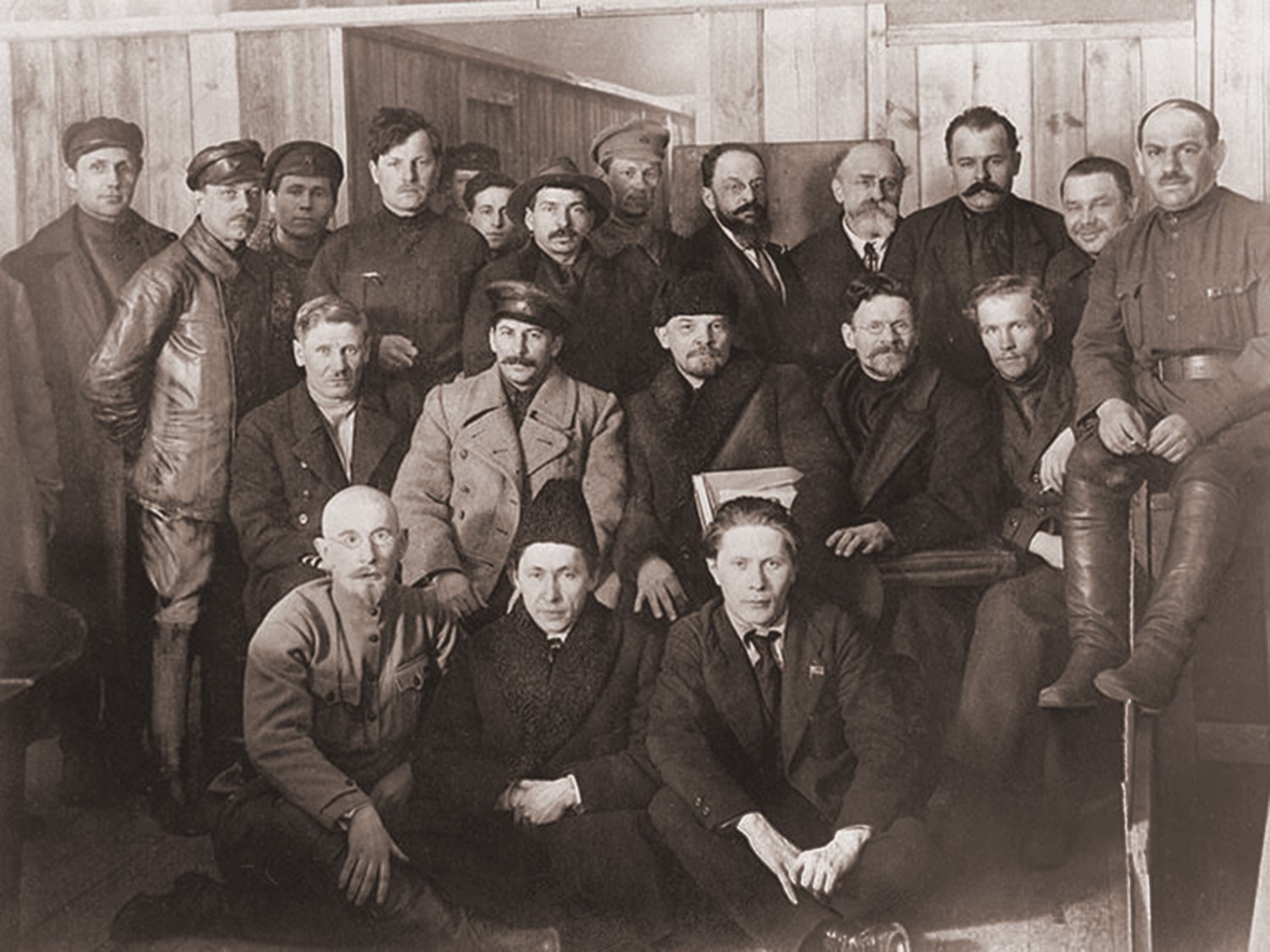 Sepia-toned photograph of participants at the 8th Congress of the Russian Communist Party in 1919. The group is assembled in a wooden room. Central figures include Stalin, Vladimir Lenin, and Mikhail Kalinin, surrounded by other delegates dressed in a mix of military and civilian attire.