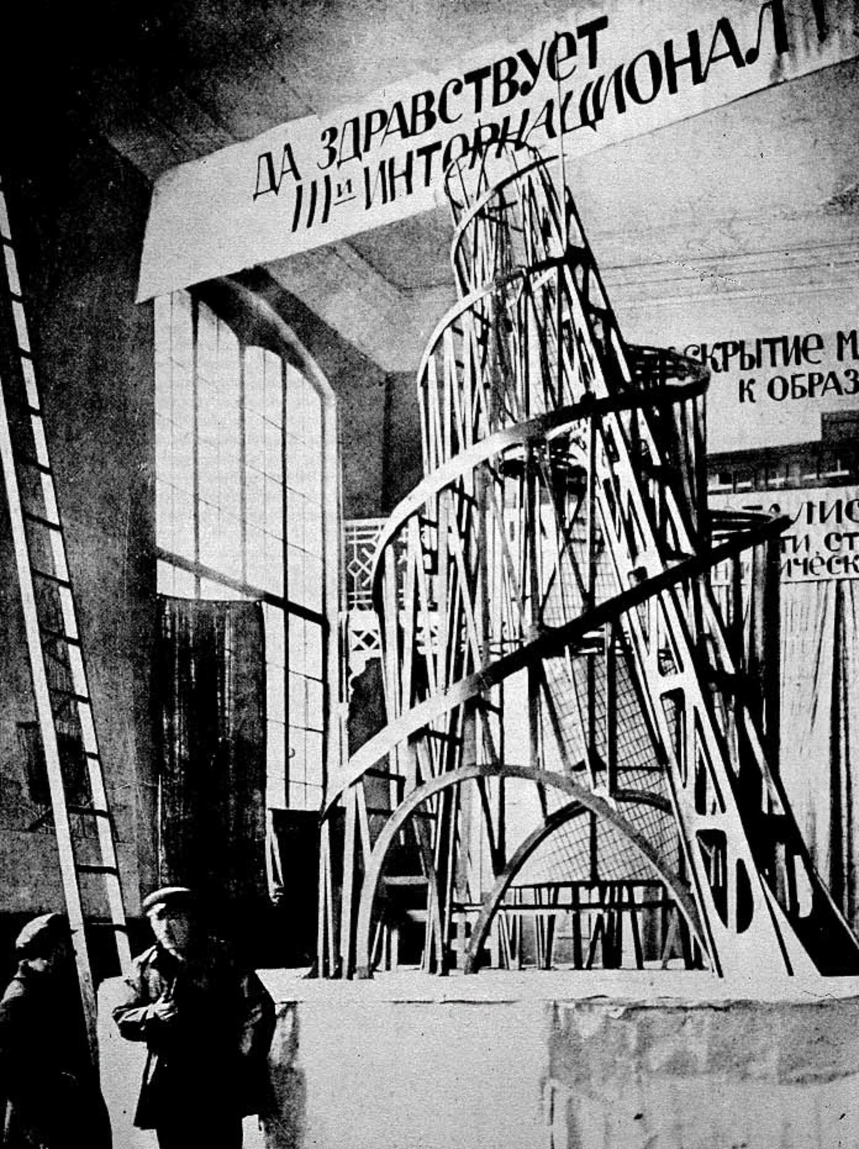 An evocative black-and-white photograph from 1919 showcases the model of 'Tatlin’s Tower', conceptualized by Vladimir Tatlin. The intricate tower structure, a fusion of spiraling frameworks and geometrical shapes, stands prominently against a backdrop of large windows in a spacious room. The banner reading 'ДА ЗДРАВСТВУЕТ III-й ИНТЕРНАЦИОНАЛ' ('LONG LIVE THE THIRD INTERNATIONAL') hangs overhead. In the foreground, Vladimir Tatlin and an assistant are seen observing the tower, providing a sense of scale to the architectural masterpiece envisioned as a symbol of modernity and progress.