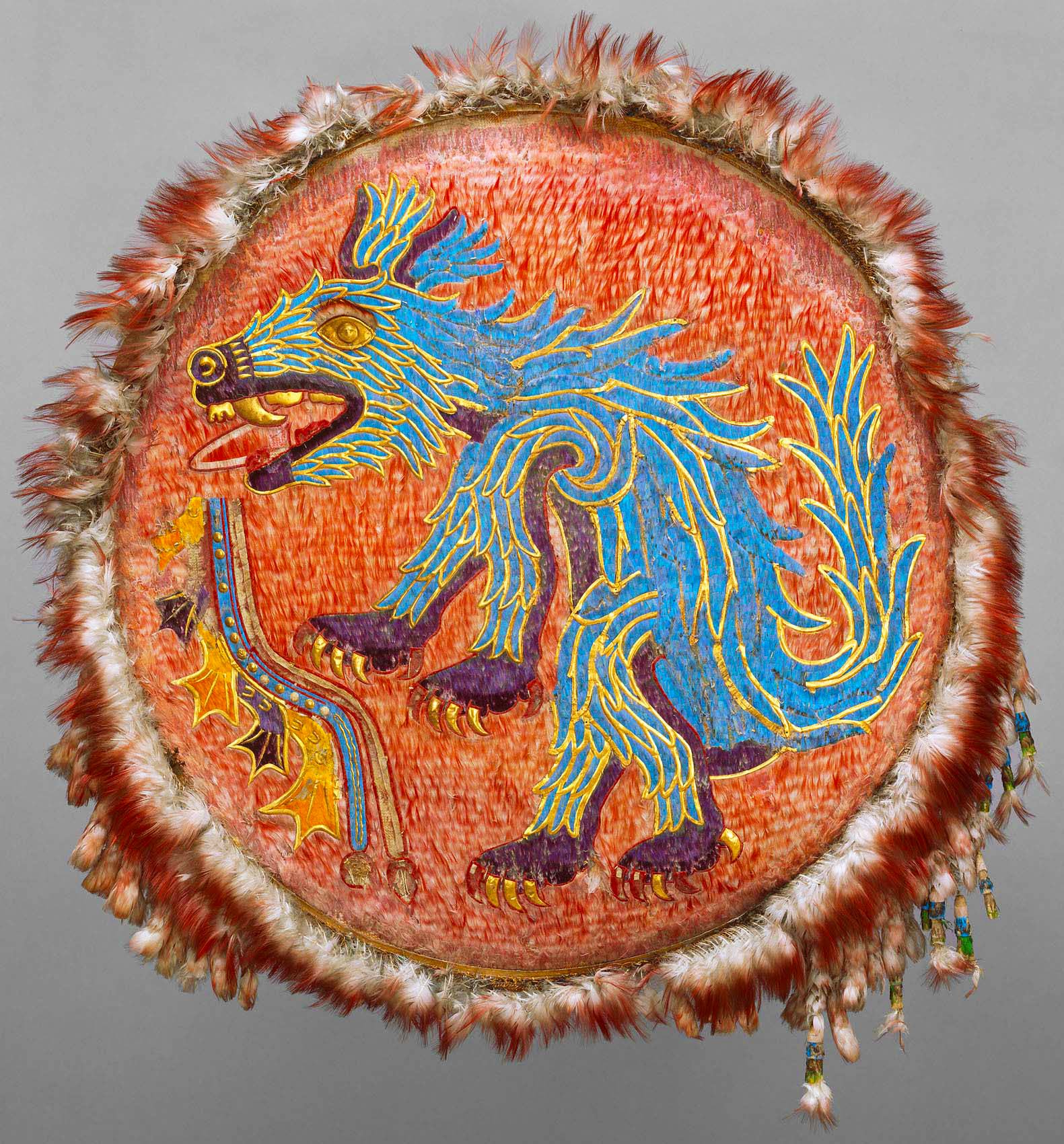 A vibrant and intricately detailed shield showcasing the image of Ahuizotl, an Aztec mythological creature. The creature, predominantly painted in bright blue, exhibits fierce features such as large, sharp claws, a snarling face with prominent fangs, and a distinct curling tail ending with a hand. The background of the shield displays a vivid orange-red texture, imitating fur or feathers, enhancing the contrast with the blue figure. The shield's perimeter is bordered with a layer of fluffy, light-colored feathers, which adds a tactile dimension to the artifact. Dangling from the bottom edge of the shield are several small adornments or talismans, adding depth and movement. 
