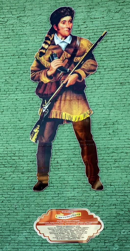 A large illustrated cut-out of American frontiersman, soldier, and politician David Crockett is mounted on a green brick wall in Lawrenceburg, Tennessee. Crockett is depicted in period attire, holding a long rifle and wearing a coonskin hat. Below the image, there's a decorative plaque that reads 'Welcome to Lawrenceburg - Home of David Crockett. Frontiersman, Justice of the Peace, Lawrence County Magistrate, and Congressman. Places of interest to visit...'. The plaque lists various attractions related to David Crockett in the area.