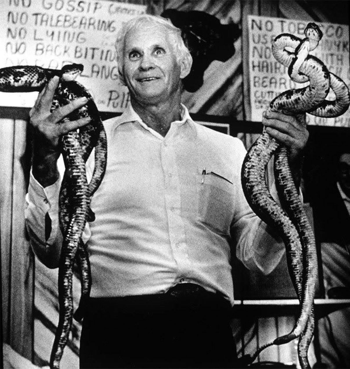 Black and white photograph of George Hensley with white hair, looking upwards while holding large serpents in his hands. Behind him, a board displays various church rules such as 'No Gossip' and 'No Tobacco'.