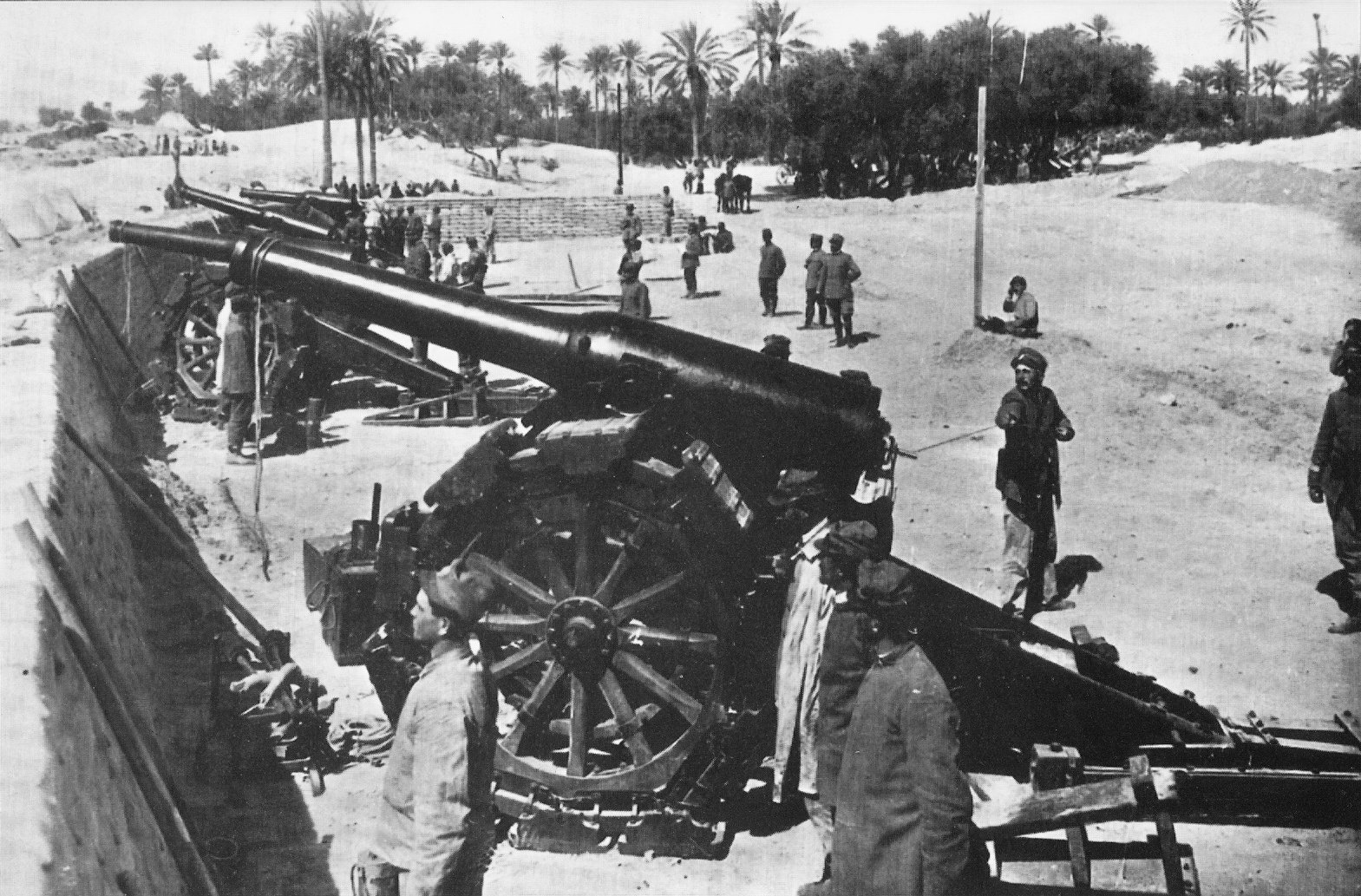 A historical photograph from the Italo-Turkish War in 1911, showcasing an Italian artillery battery positioned near Tripoli. The massive cannon, supported by large wheels and mechanisms, is prominent in the foreground. Soldiers in period uniforms are dispersed around the area, with some attending to the artillery, while others stand or move about, conducting various duties. The background is dotted with palm trees and sparse structures.