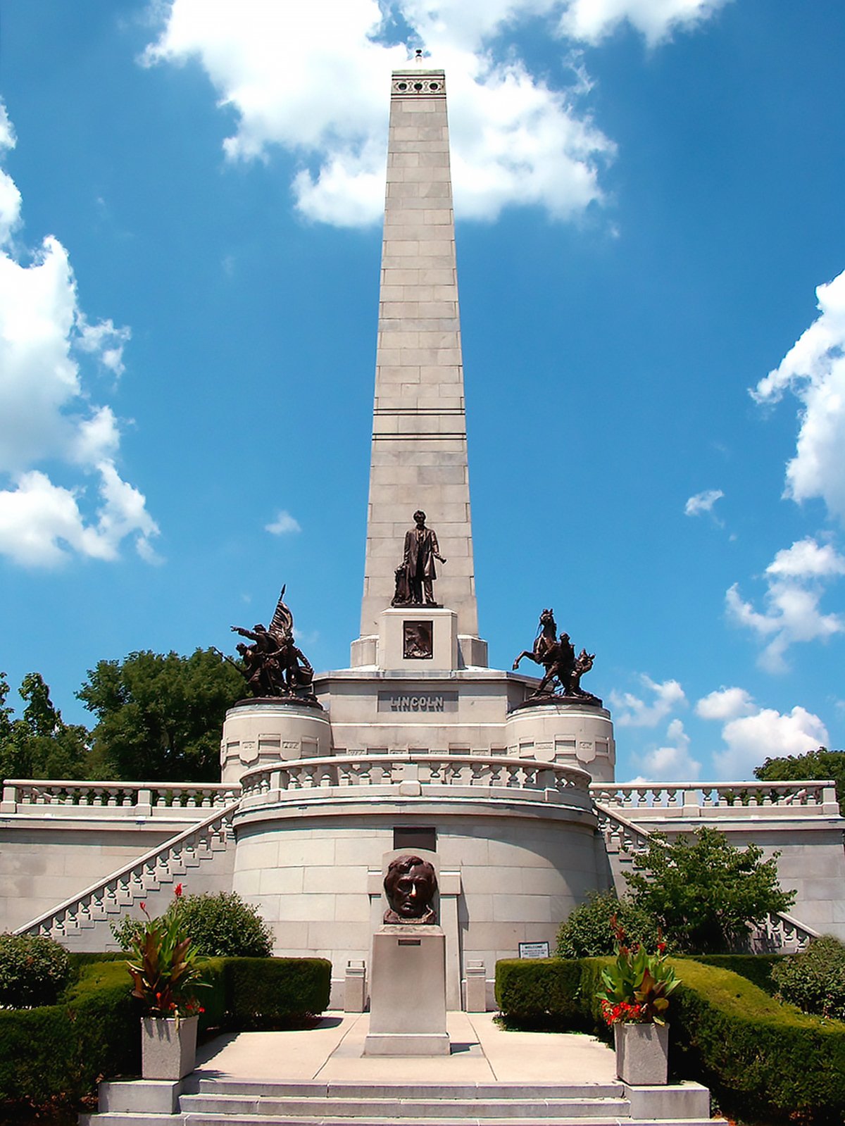 This is an image of the Abraham Lincoln Tomb in Springfield, Illinois. The towering monument, topped by an obelisk, is surrounded by several bronze statues, with a bust of Lincoln placed prominently at the front. Stairs lead up to the structure set against a backdrop of a clear blue sky with a few scattered clouds. The monument serves as a memorial to the 16th President of the United States.