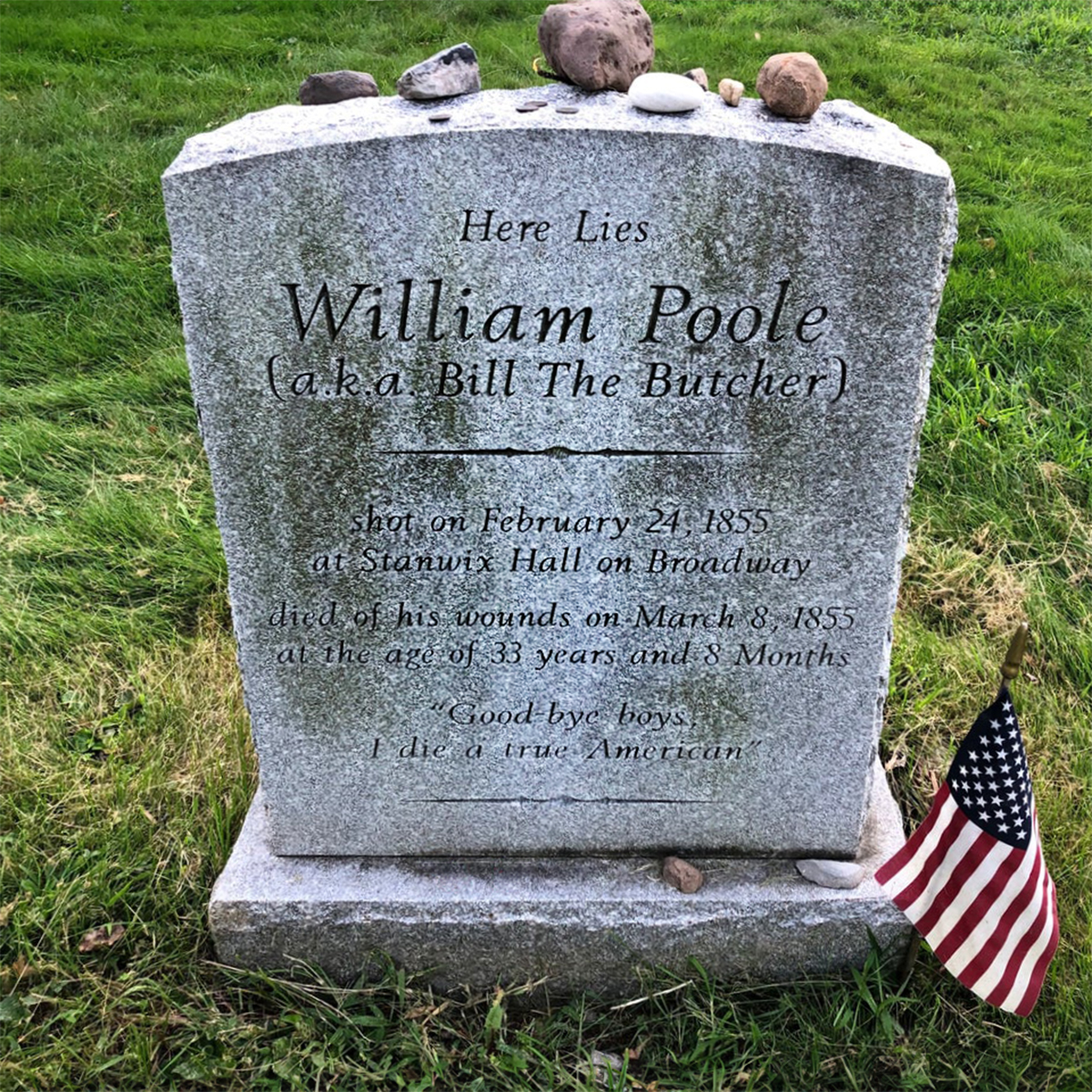 This image shows the gravestone of William Poole, also known as Bill The Butcher. The inscription reads that he was shot on February 24, 1855, at Stanwix Hall on Broadway, and died of his wounds on March 8, 1855, at the age of 33 years and 8 months. The epitaph says "Good bye boys I die a true American." Small stones are placed on top of the gravestone, which is a tradition often associated with respect or remembrance. An American flag is planted in the ground next to the grave.
