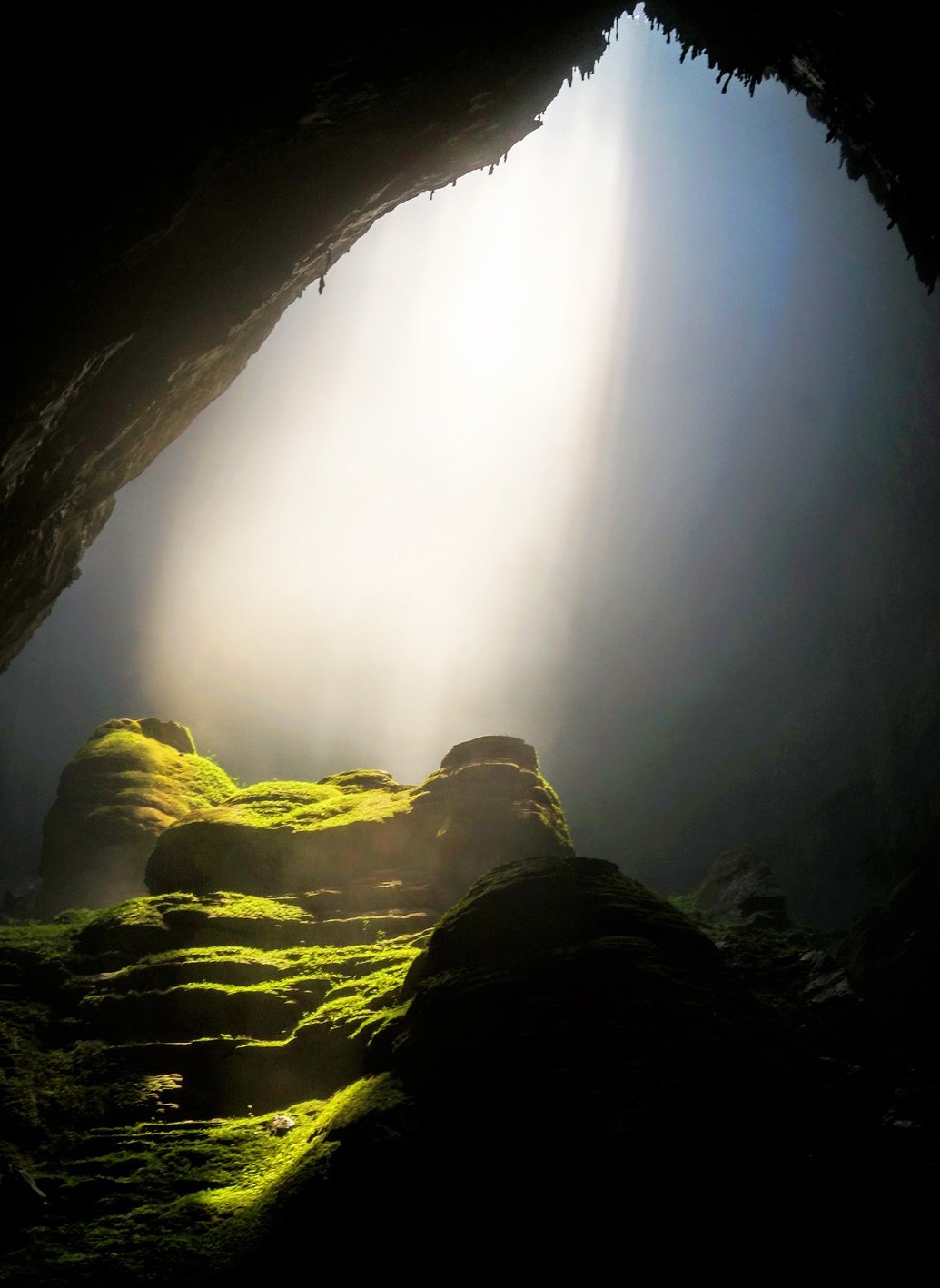 Shafts of light piercing through the opening of a cave onto moss-covered rocks, symbolizing the Druidic belief in the soul's immortal journey to the Otherworld.