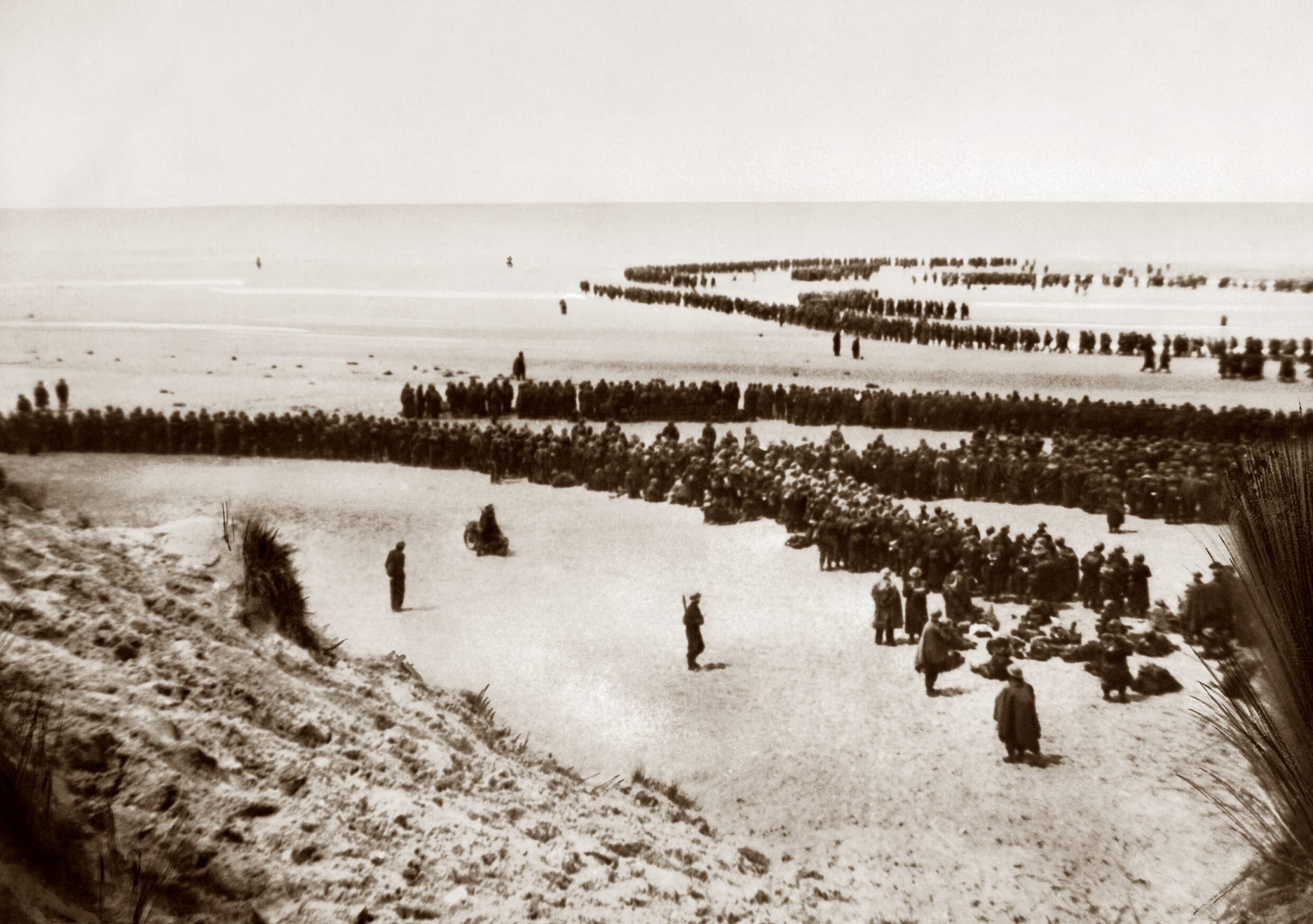 Historical black and white photo showing British troops in long, orderly lines on the sands of Dunkirk beach, waiting for evacuation between 26-29 May 1940. Some soldiers are seated on the ground, while others stand in formations, with the vast, open beach and horizon in the background. The scene is calm, despite the urgent circumstances.